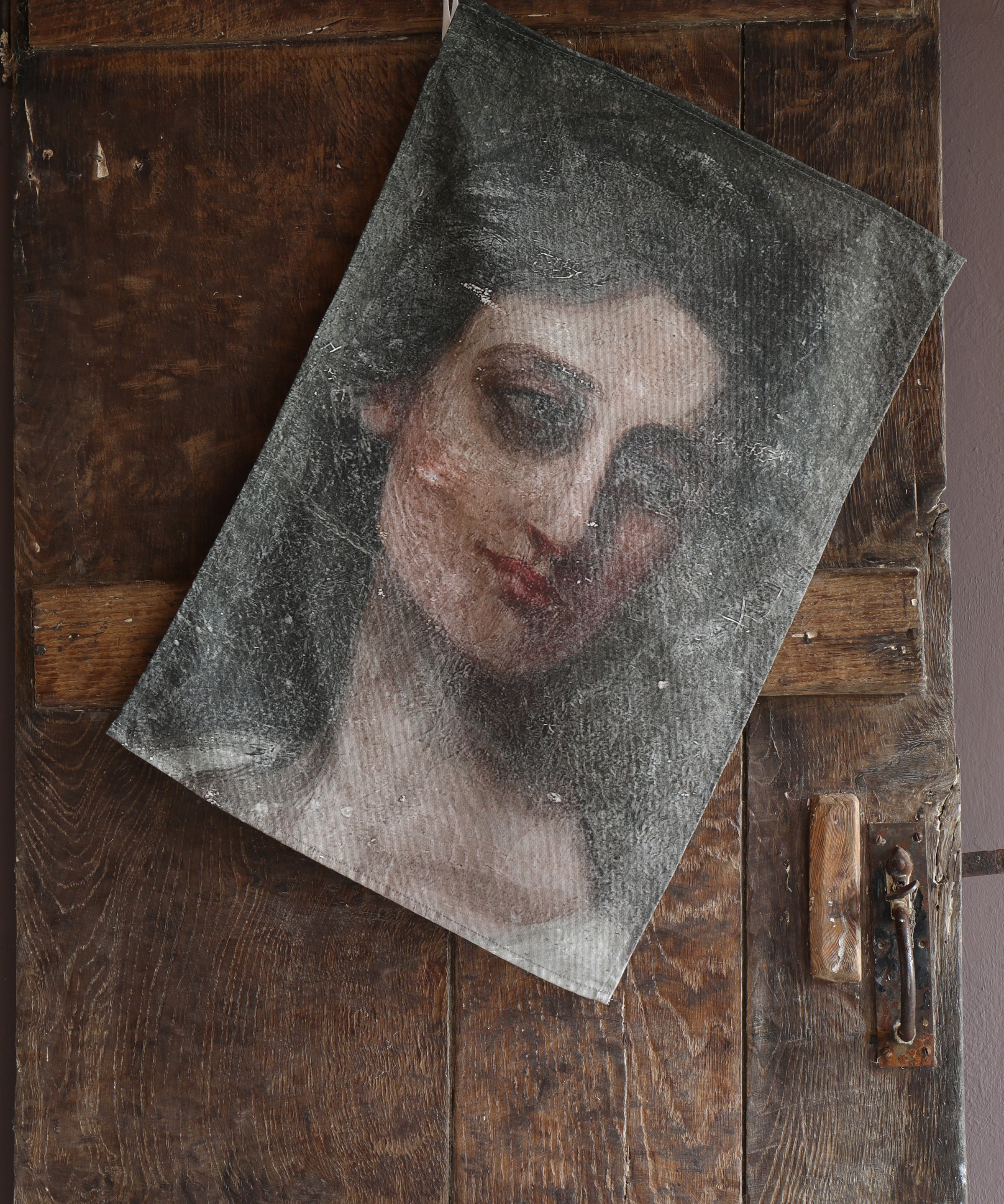 Tea towel hanging on an old wooden door with an image of a woman from an oil painting looking wistfully to the side.