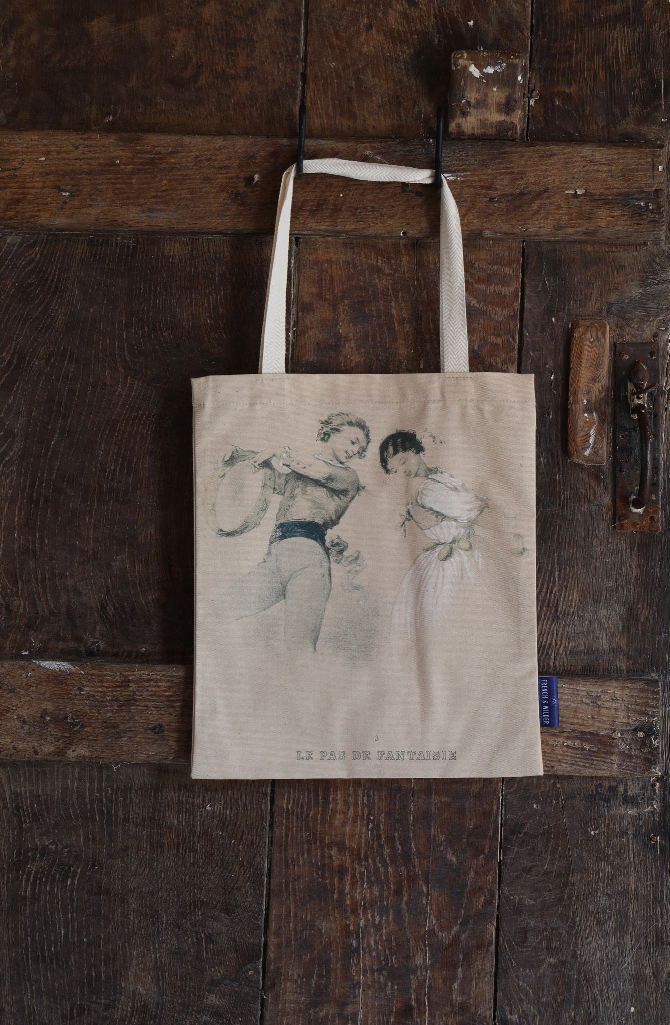 Tote bag with a vintage line-drawn image featuring two young lovers with the text 'Le Pas de Fantaisie' hanging next to a dark wooden door