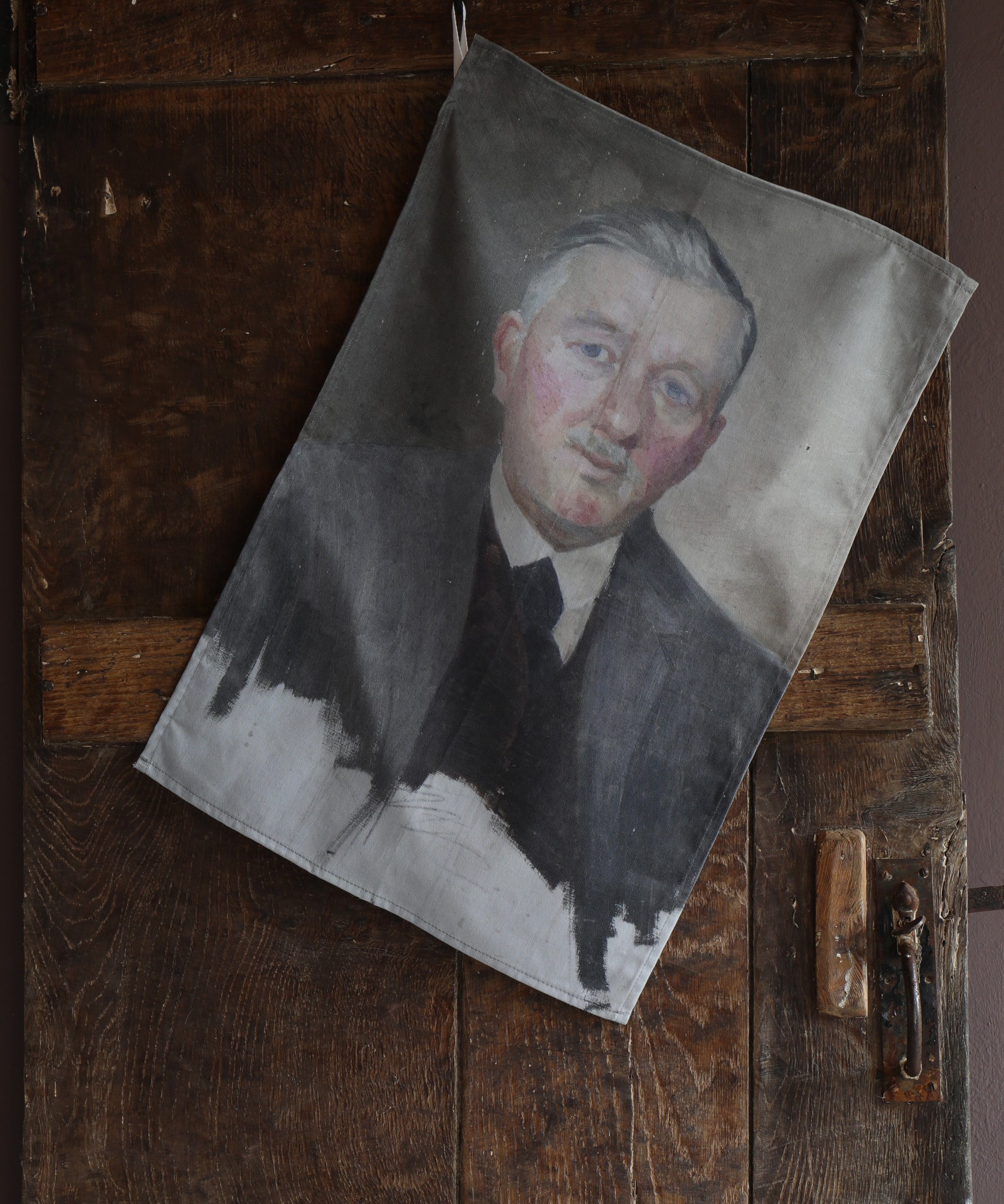 Tea towel hanging on an old wooden door with the image of a vintage half-finished portrait of smartly dressed red-faced older man. 