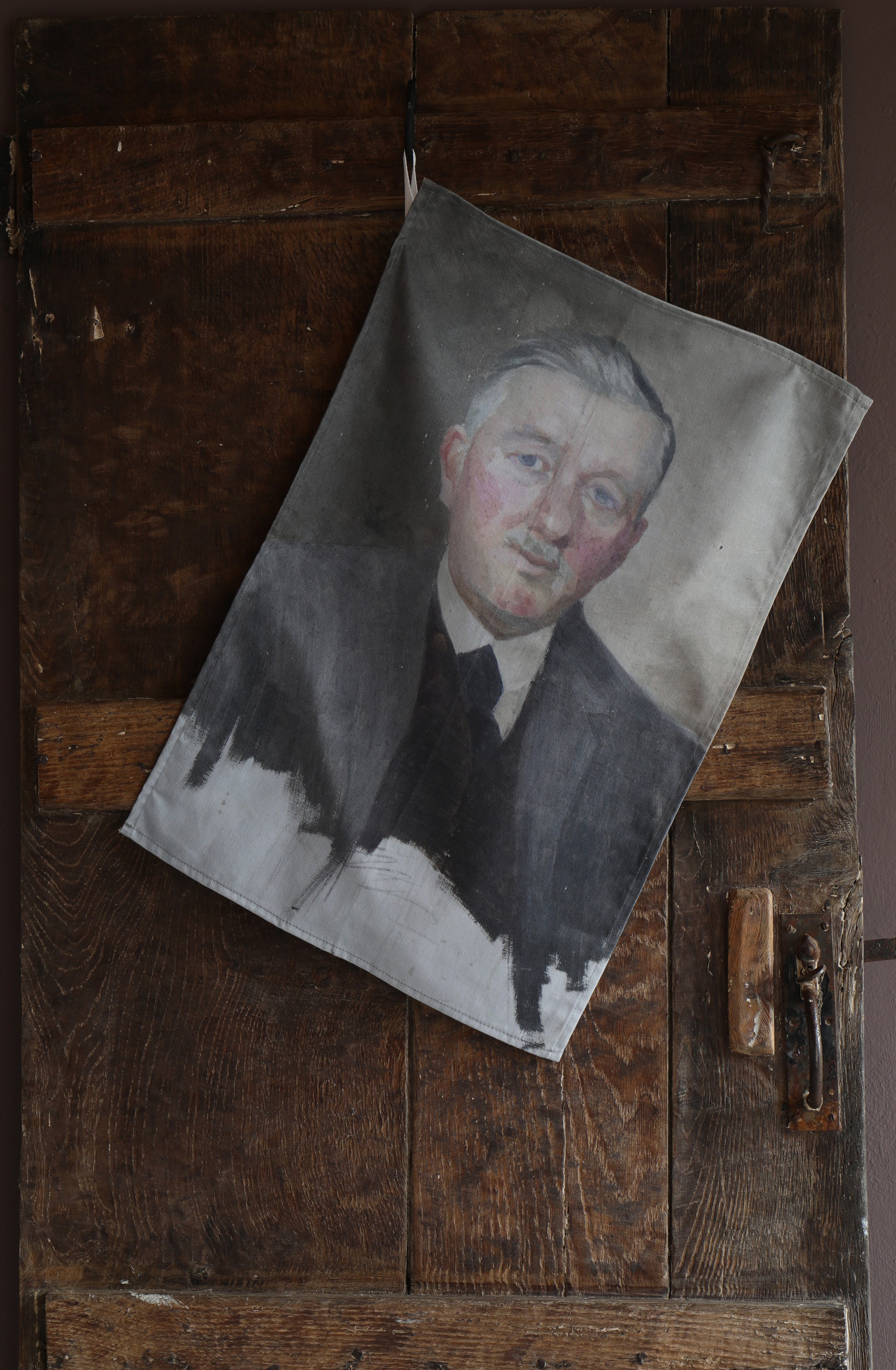 Tea towel hanging on an old wooden door with the image of a vintage half-finished portrait of smartly dressed red-faced older man. 