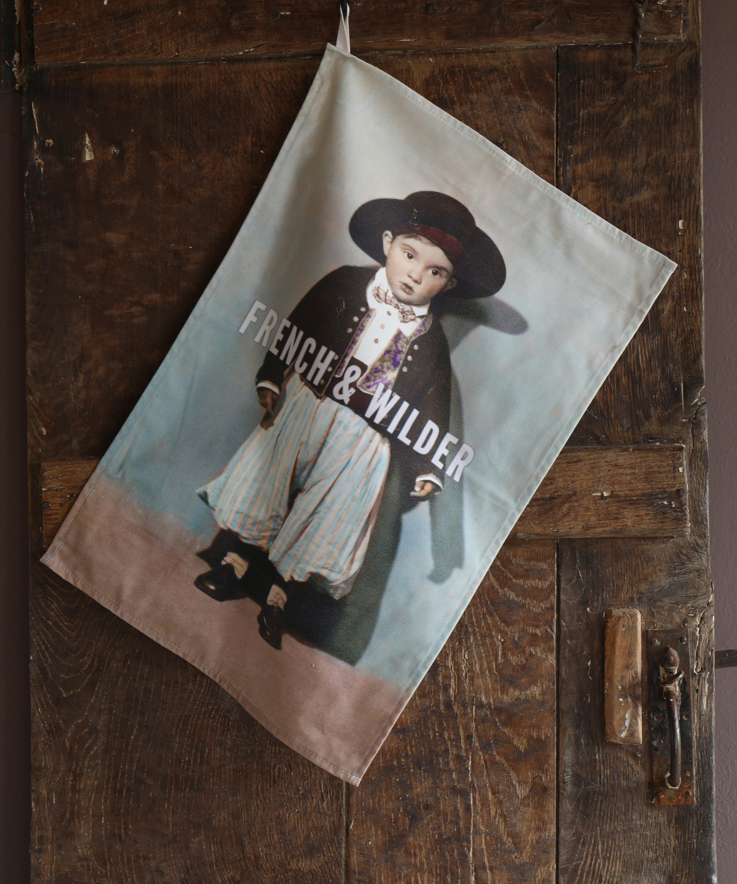 Tea towel with a printed design of a child wearing early 20th century Mediterranean costume of baggy trousers, patterned waistcoat, jacket and hat, featuring the brand 'French & Wilder', hanging on a wooden door.