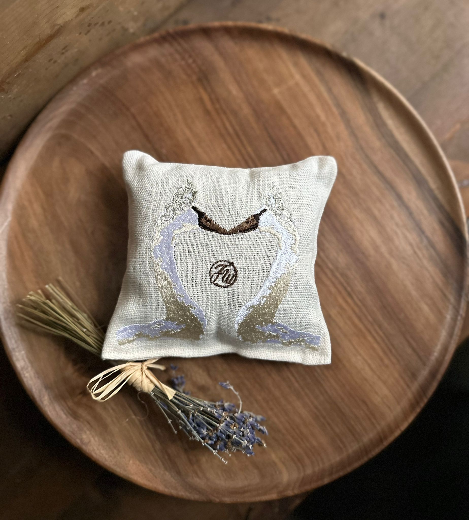Decorative lavender pillow featuring an embroidered design of two swans facing each other placed on a wooden plate with a small bunch of lavender adjacent