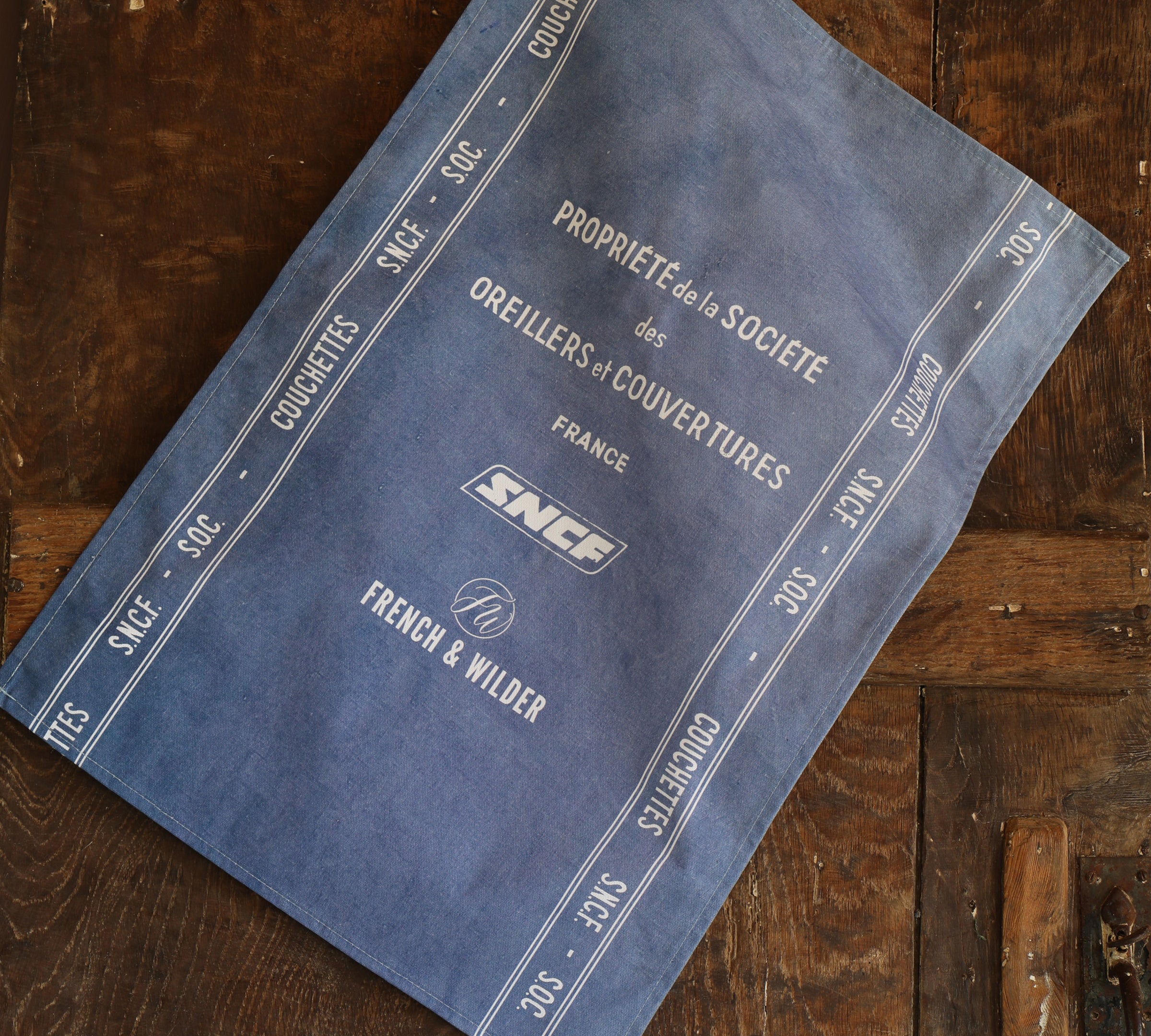 Blue fabric tea towel featuring white SNCF logos and text hanging next to a dark wooden surface