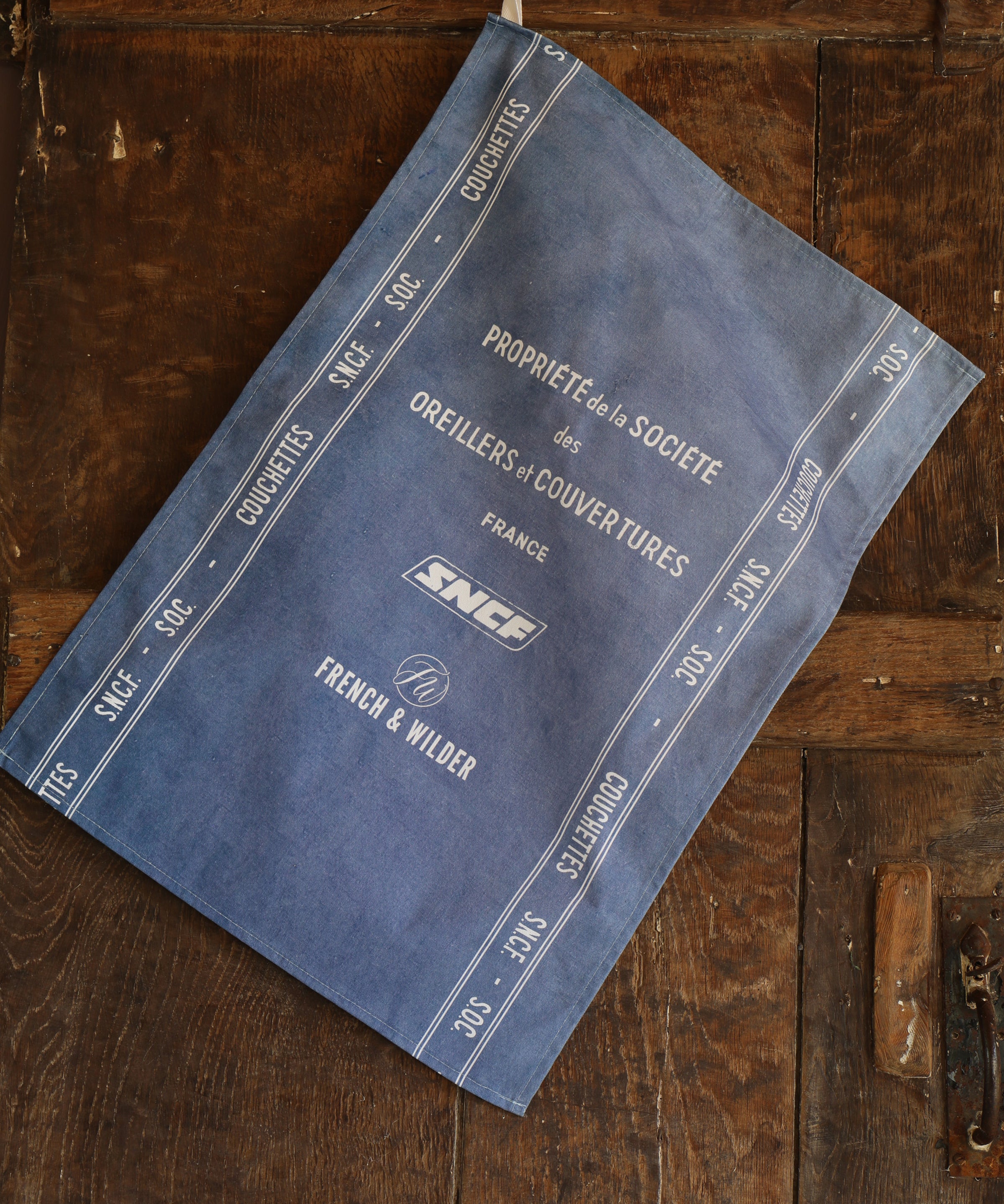 Blue fabric tea towel featuring white SNCF logos and text hanging next to a dark wooden surface