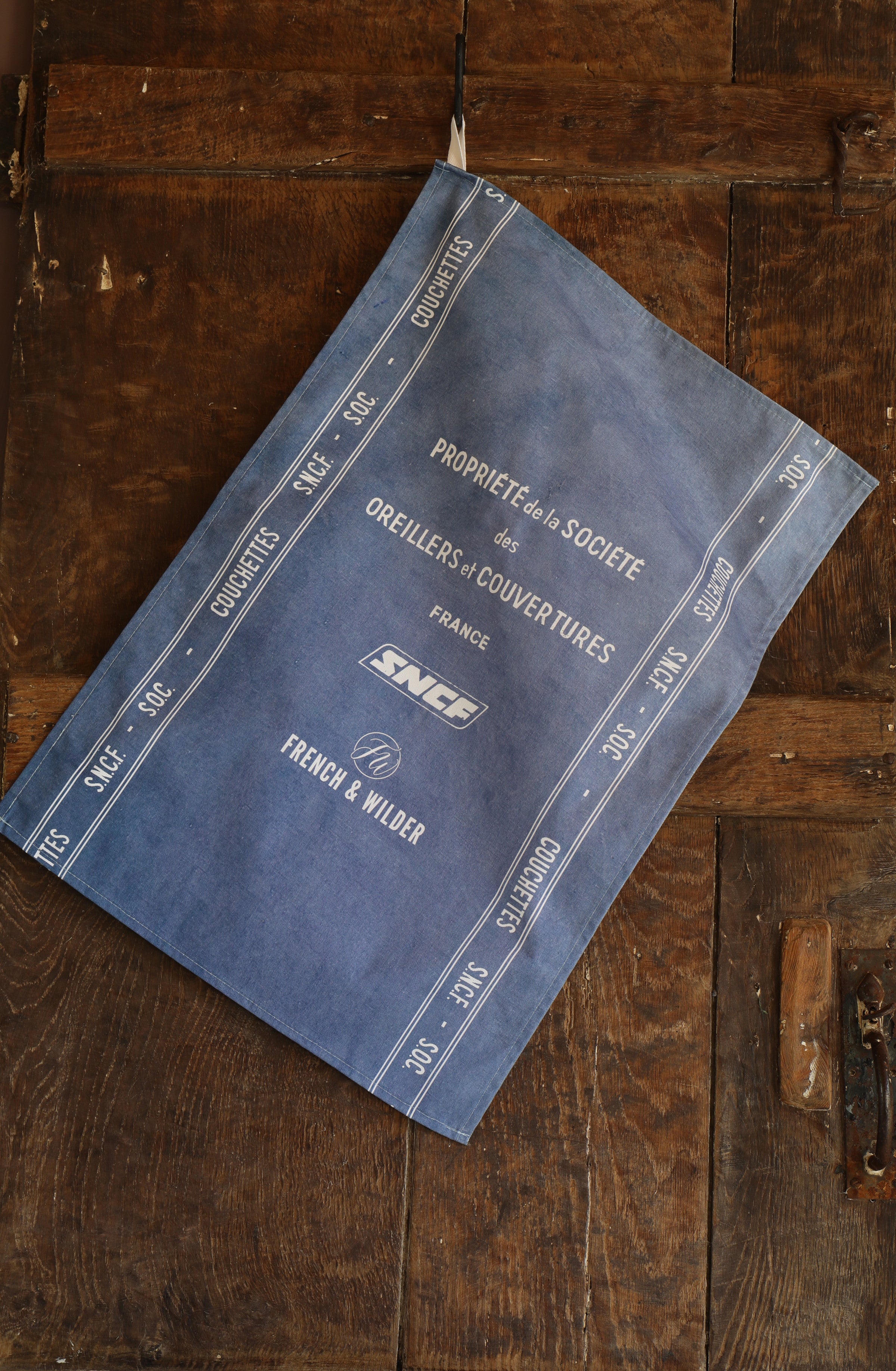 Blue fabric tea towel featuring white SNCF logos and text hanging next to a dark wooden surface