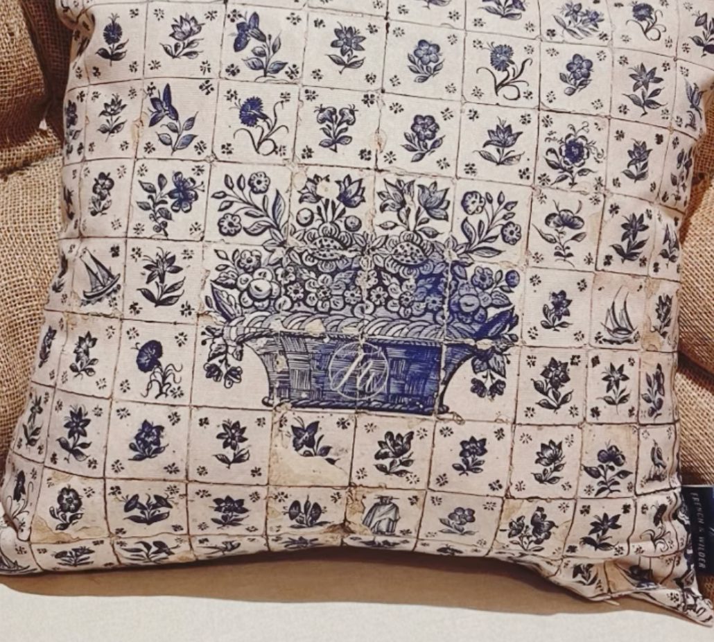 Decorative cushion featuring a blue and white tiled motif of flowers and baskets on sitting on a burlap fabric background