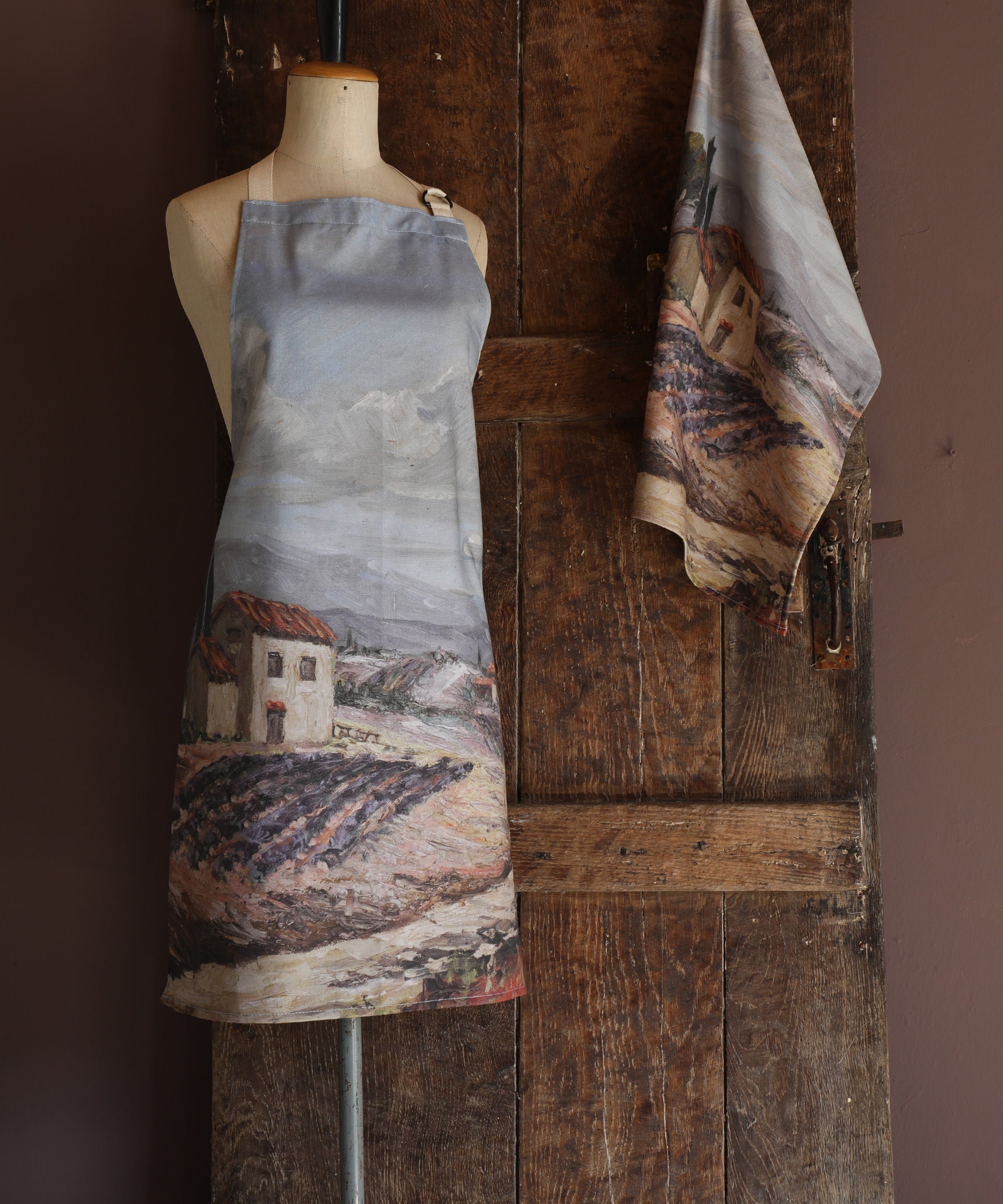 Apron with rustic Provence landscape painting design on a mannequin against a wooden wall.