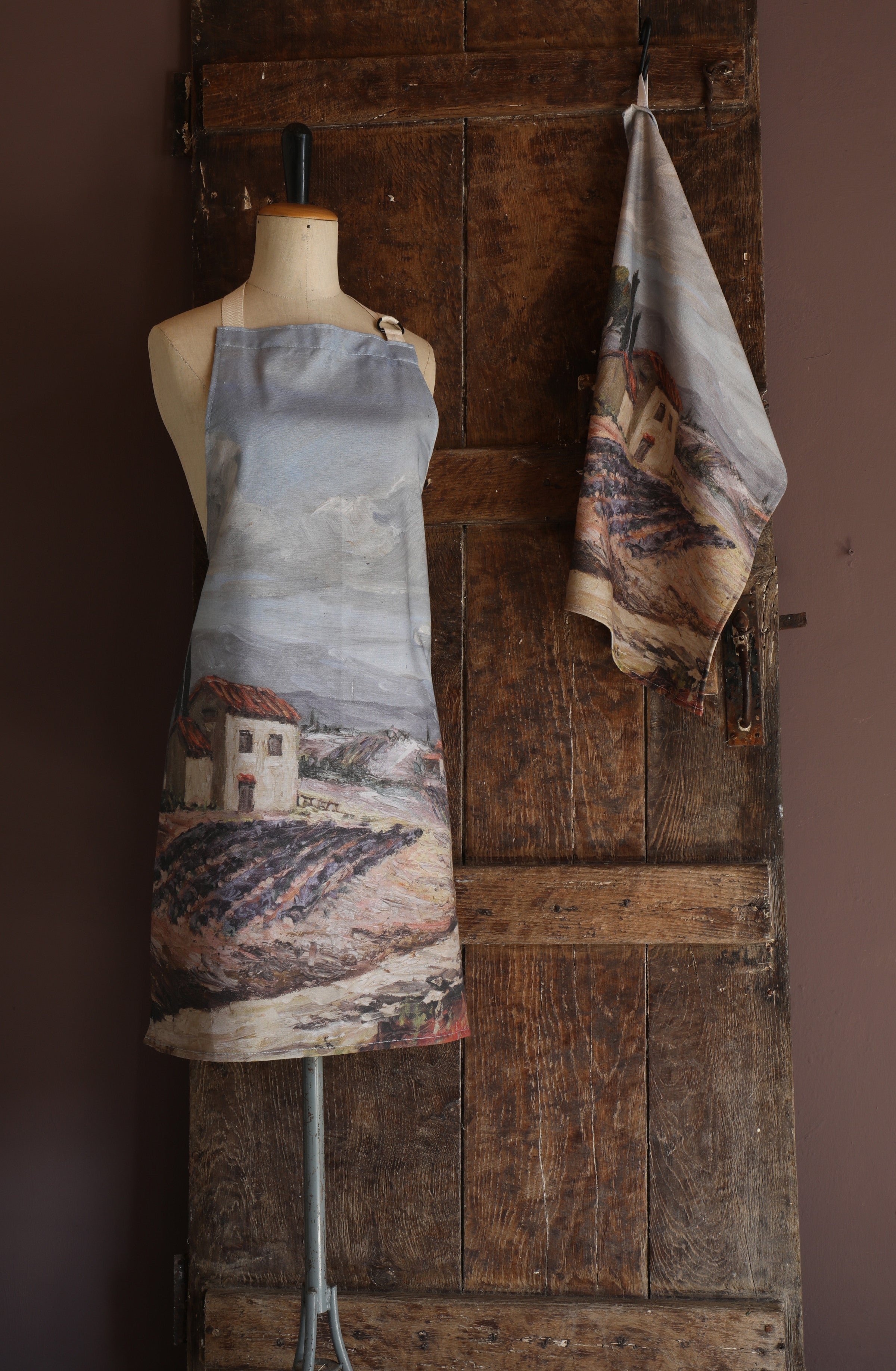 Apron with rustic Provence landscape painting design on a mannequin against a wooden wall.