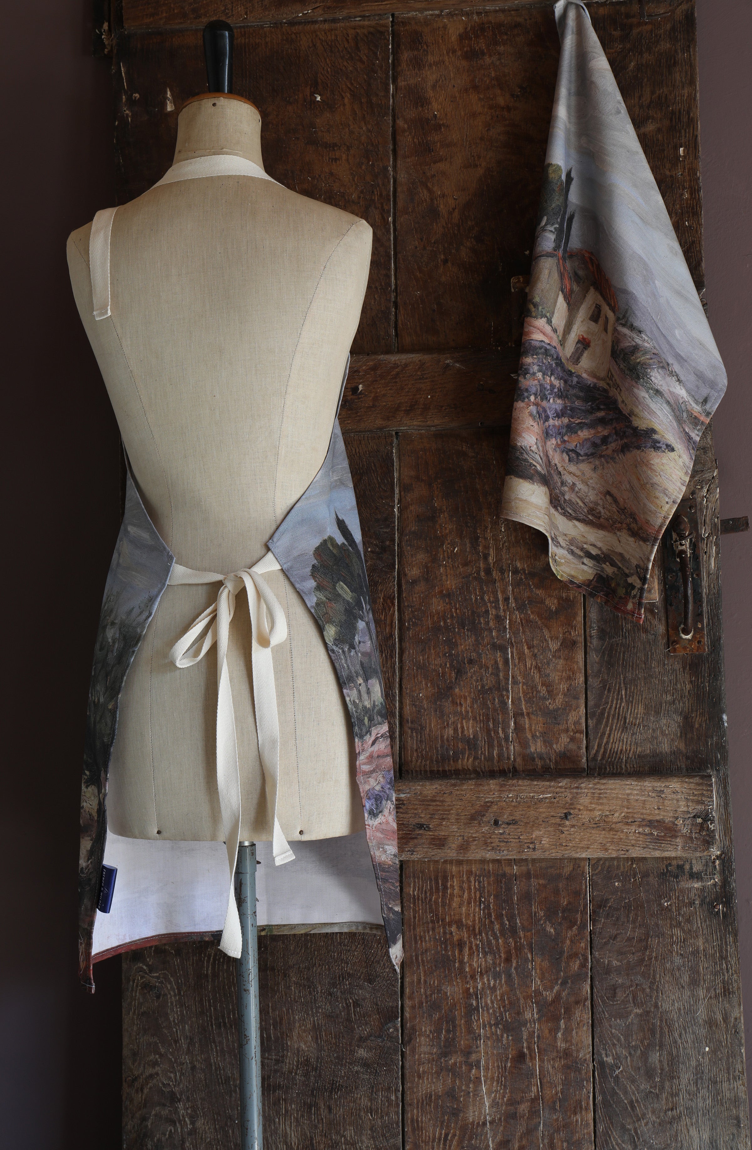 Mannequin wearing an apron with a design taken from an oil painting of a Provence scene against a wooden wall.