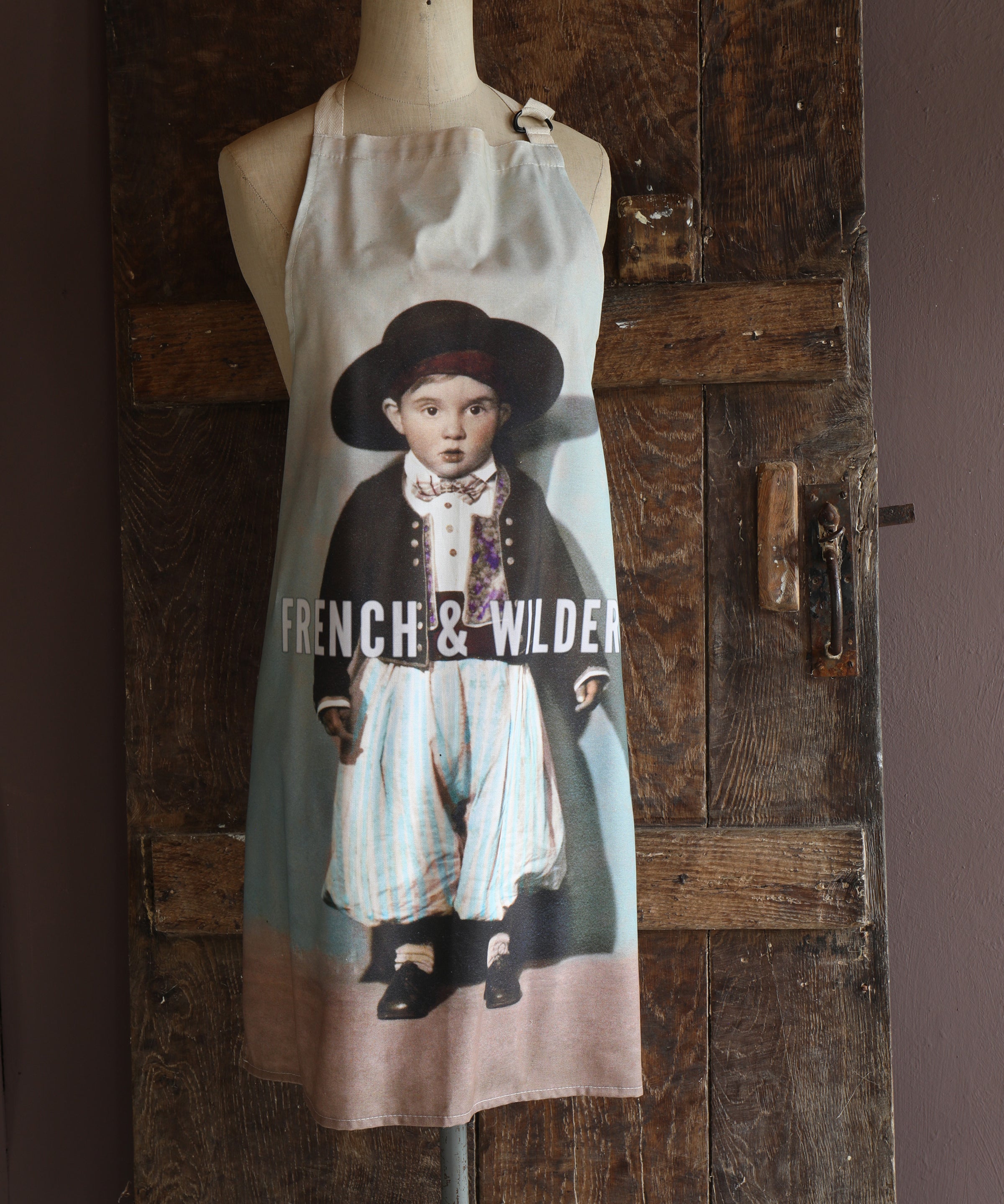 Mannequin wearing an apron with a printed design of a little boy in early 20th century traditional Mediterranean dress set against a wooden wall.