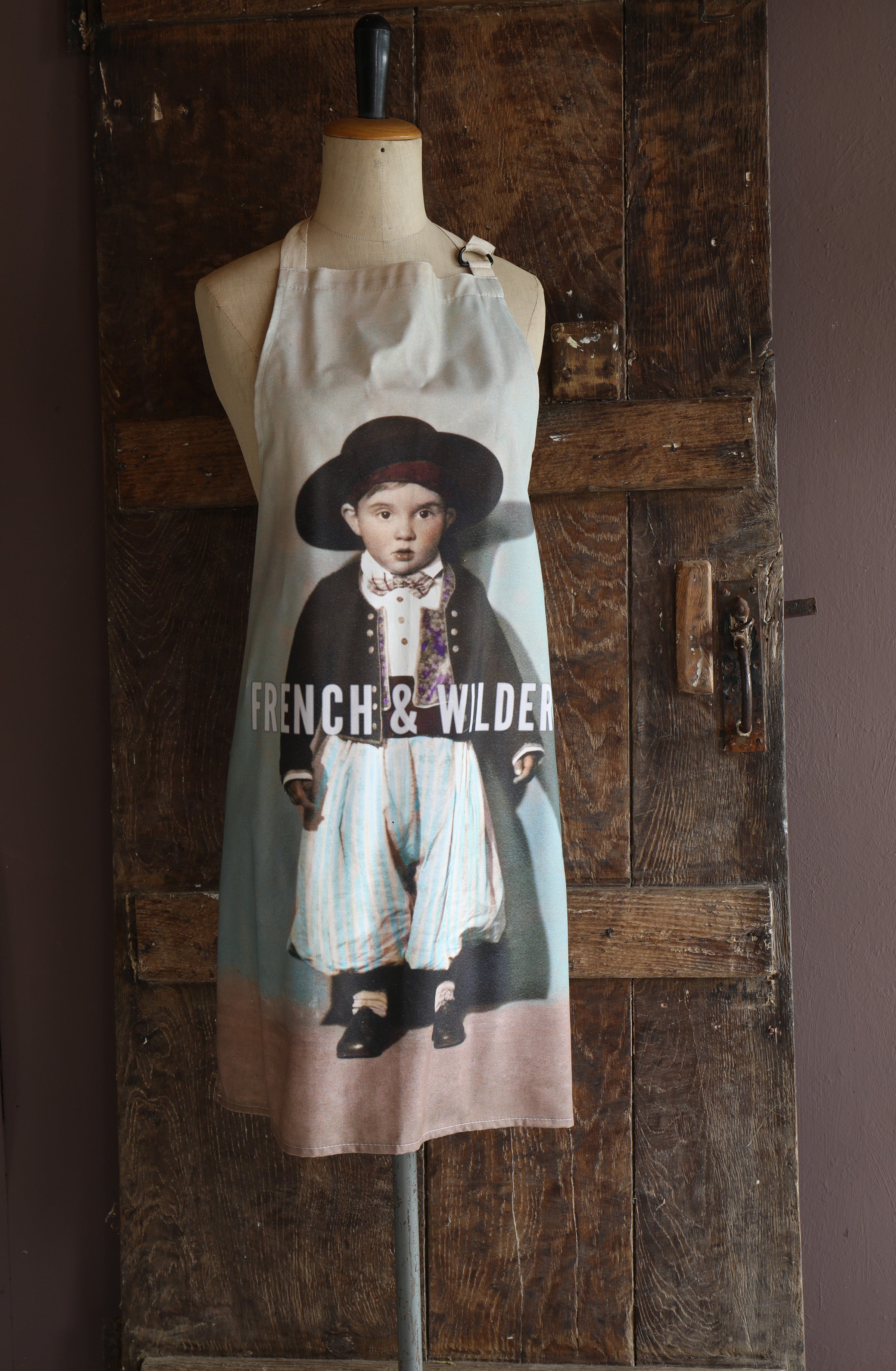 Mannequin wearing an apron with a printed design of a little boy in early 20th century traditional Mediterranean dress set against a wooden wall.