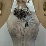 Printed apron featuring the image of a vintage mannequin wearing a decorative necklace with feathers and beads against a painted wooden background