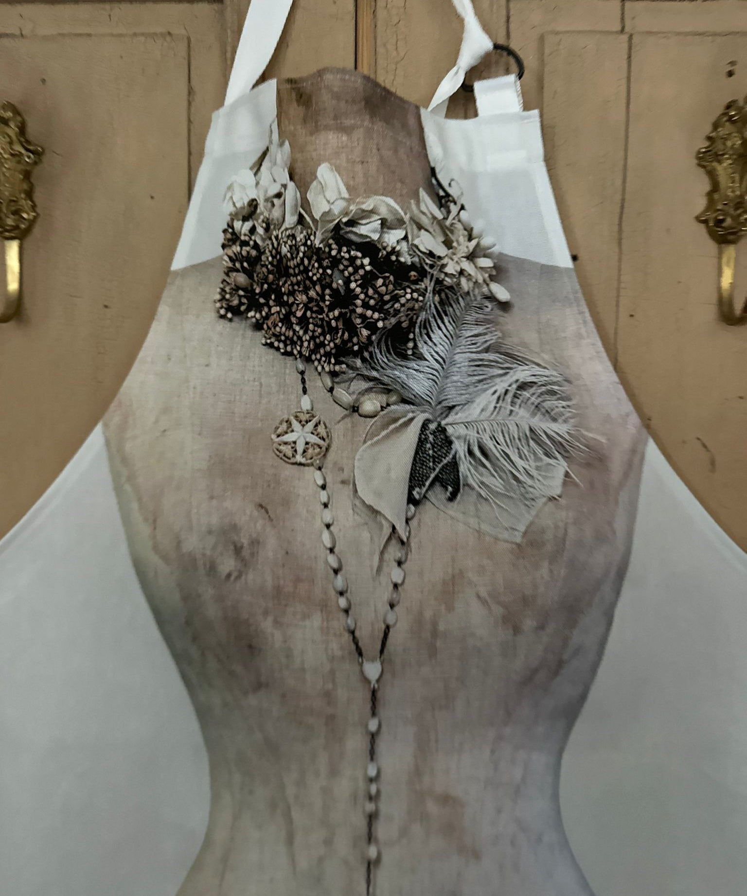 Printed apron featuring the image of a vintage mannequin wearing a decorative necklace with feathers and beads against a painted wooden background
