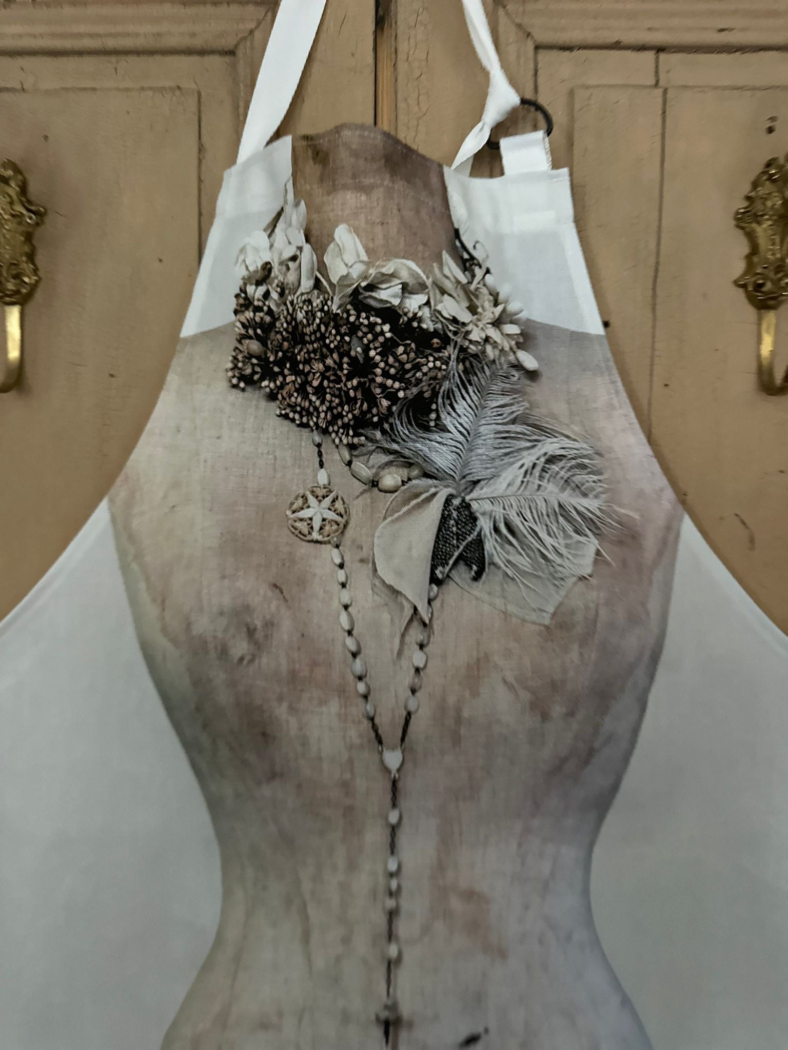 Printed apron featuring the image of a vintage mannequin wearing a decorative necklace with feathers and beads against a painted wooden background