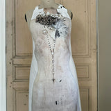 Apron featuring an antique mannequin design wearing brooches, necklaces and feathers, placed against a painted wooden door background
