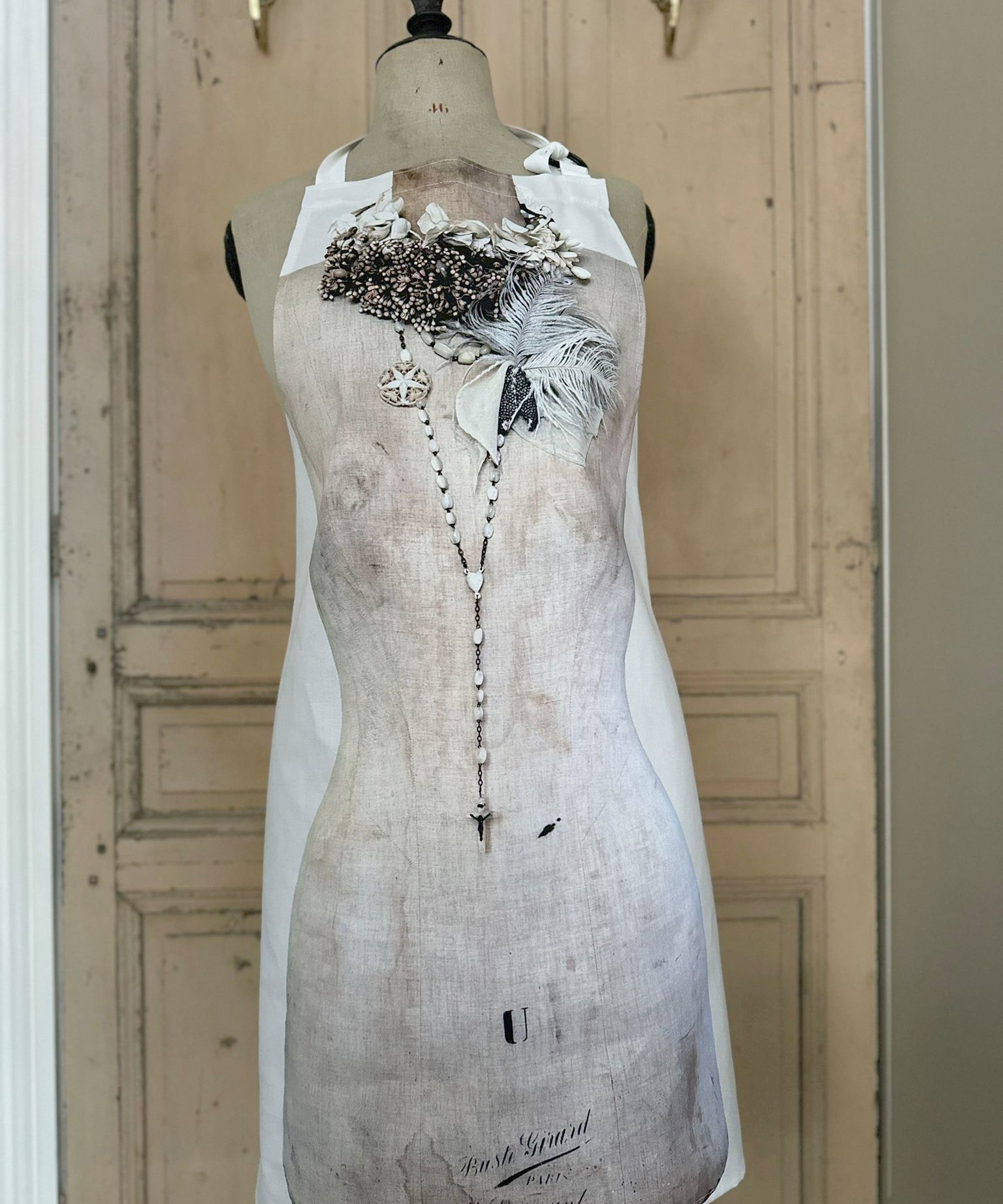 Apron featuring an antique mannequin design wearing brooches, necklaces and feathers, placed against a painted wooden door background