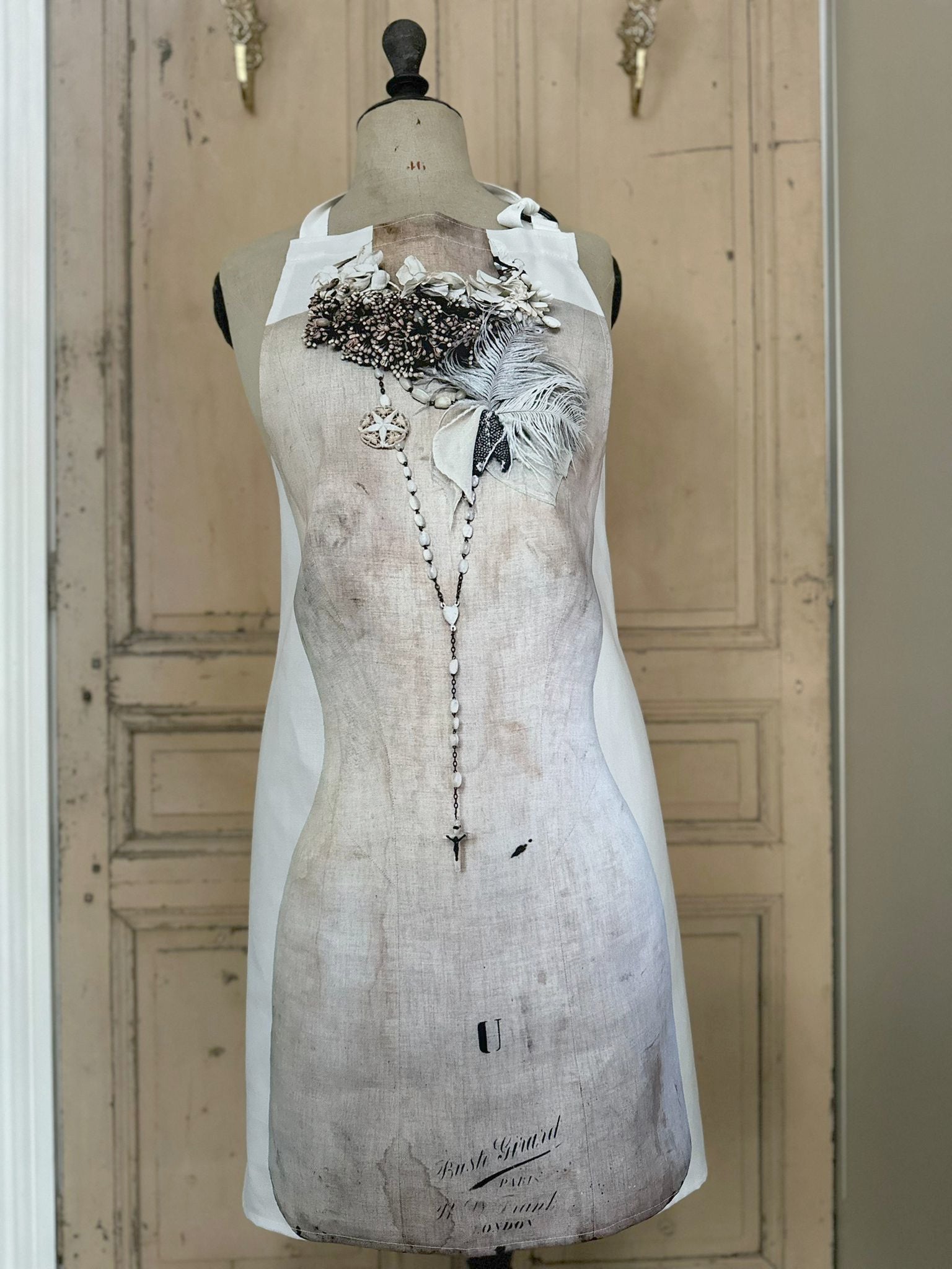 Apron featuring an antique mannequin design wearing brooches, necklaces and feathers, placed against a painted wooden door background