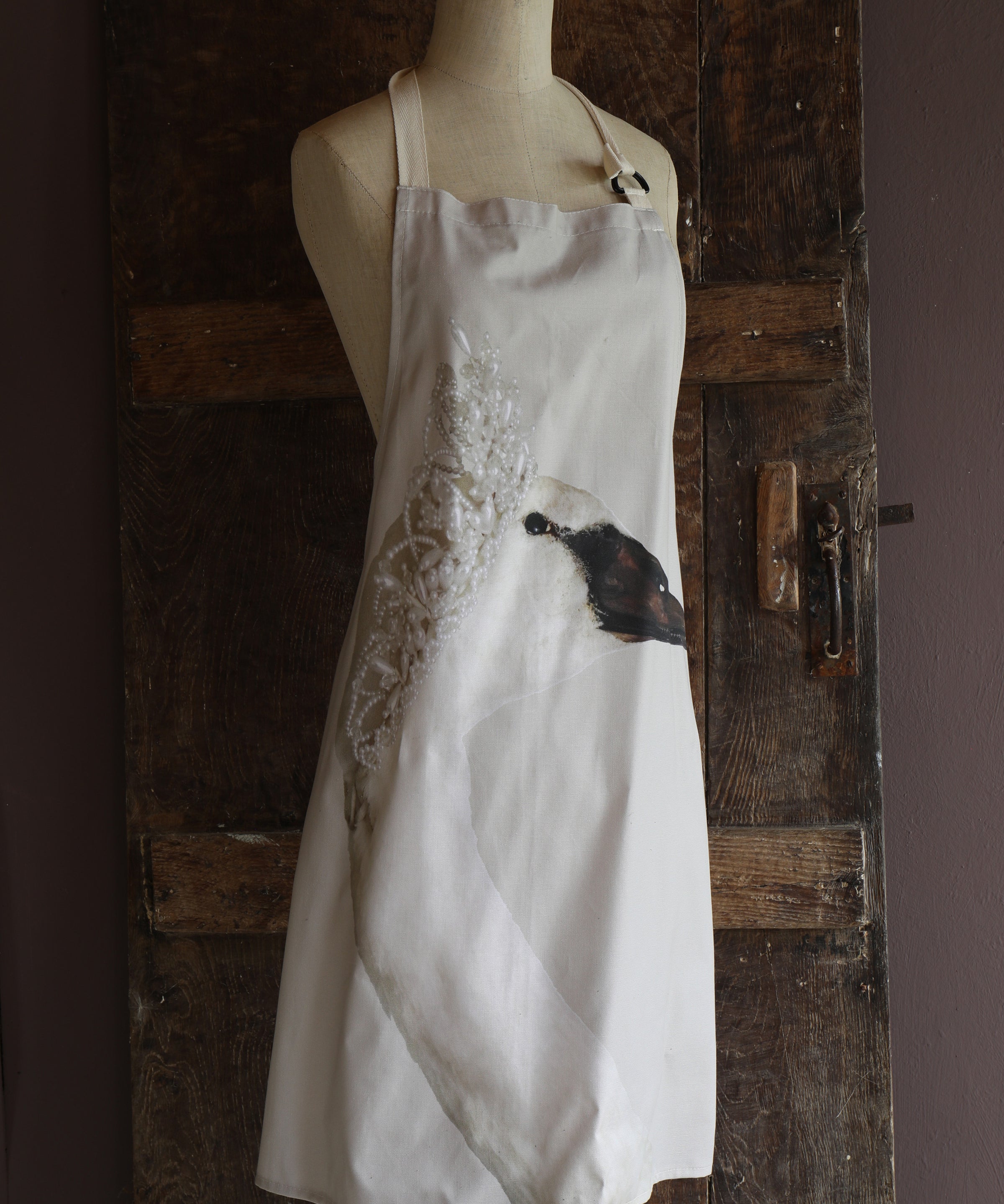 Apron with a motif of a swan wearing a tiara placed on a mannequin against a rustic wooden door