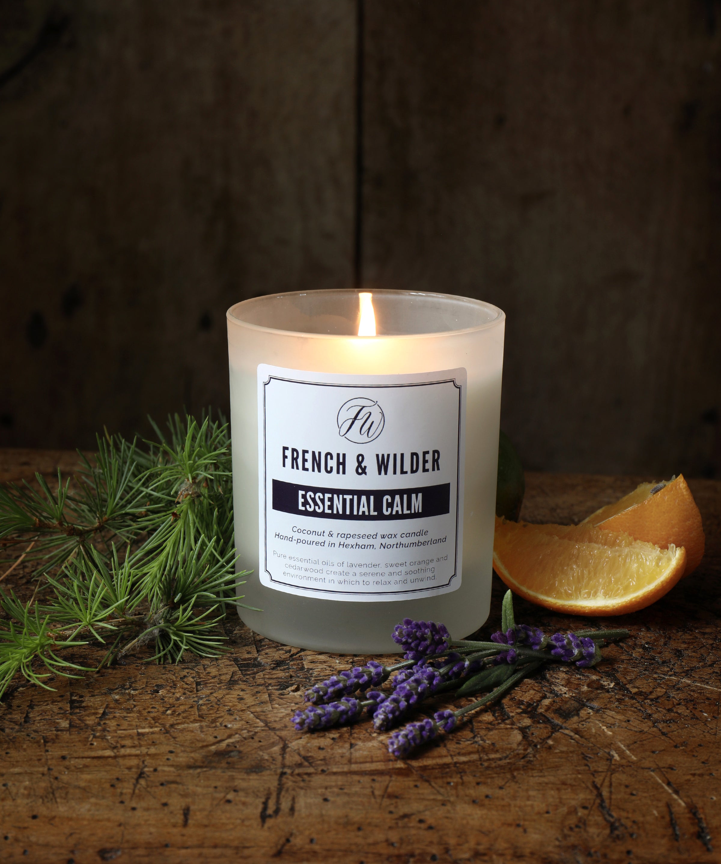 Candle in a frosted glass container on a wooden surface with lavender, pine branch and an orange slice