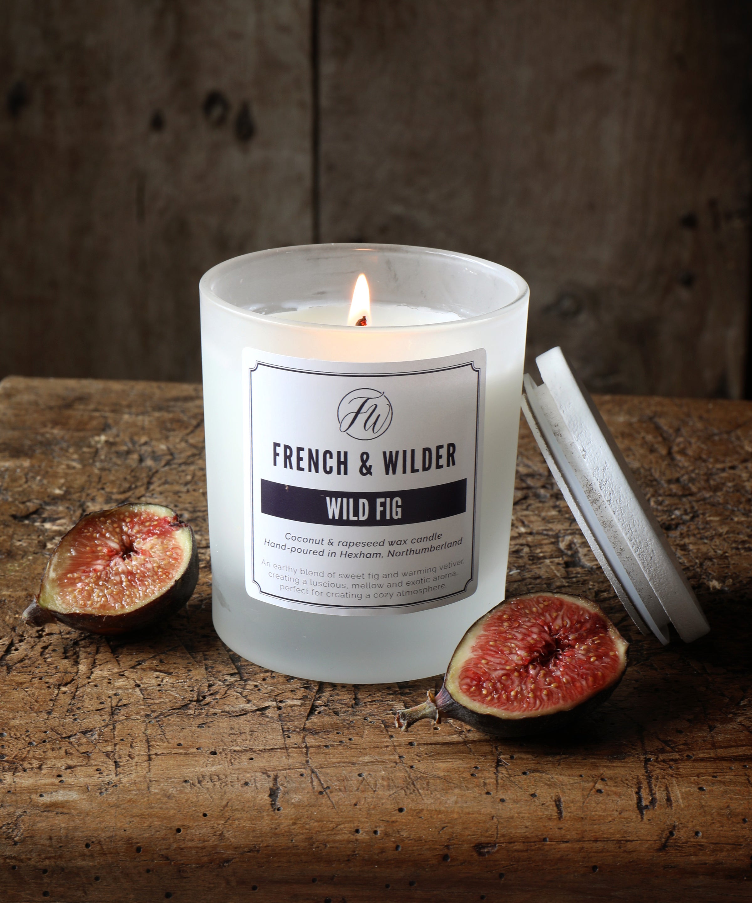 Candle in a frosted white glass container on a wooden surface next to figs