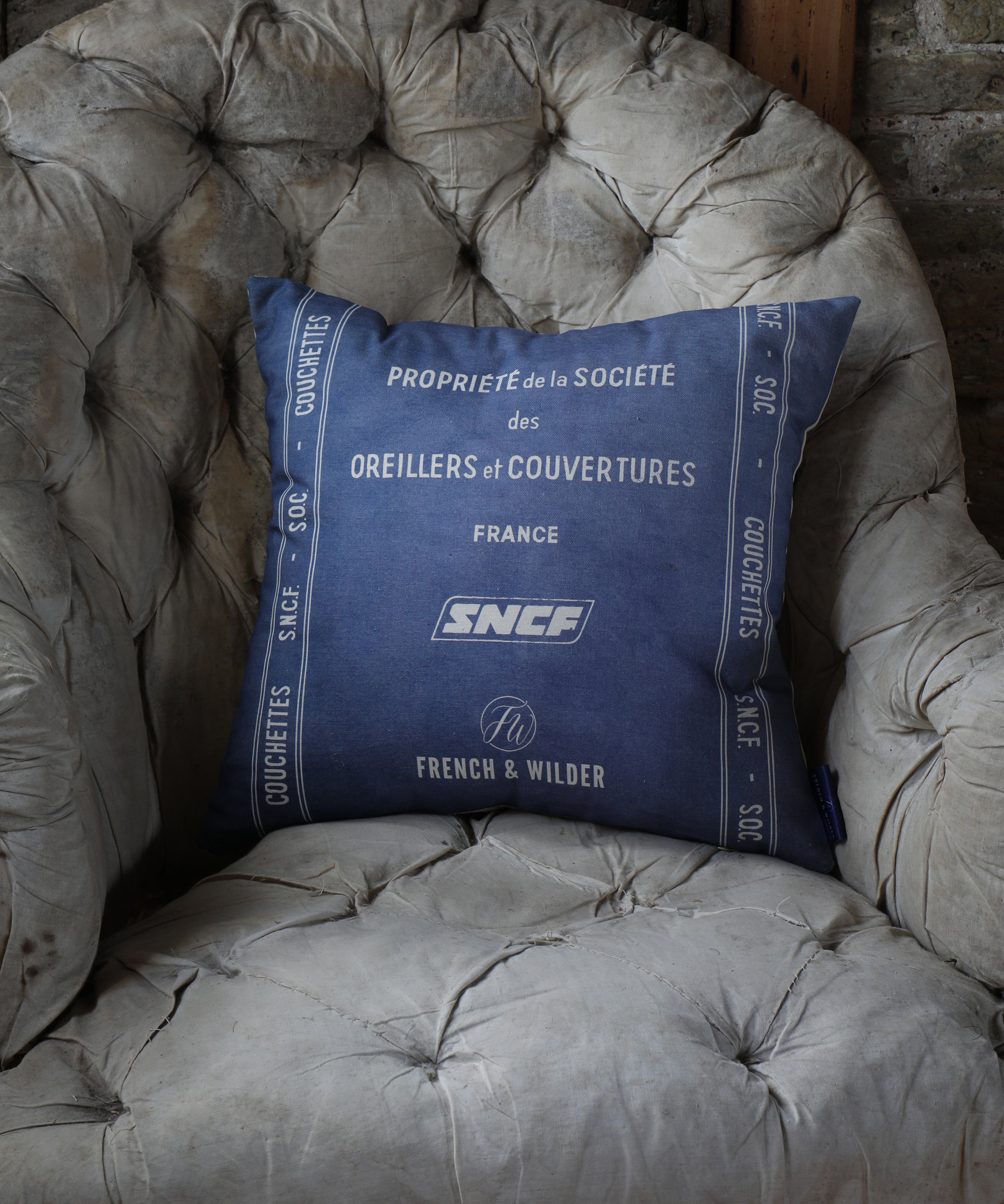 Cushion with blue and white SNCF design placed on a shabby vintage chair against a brick wall.