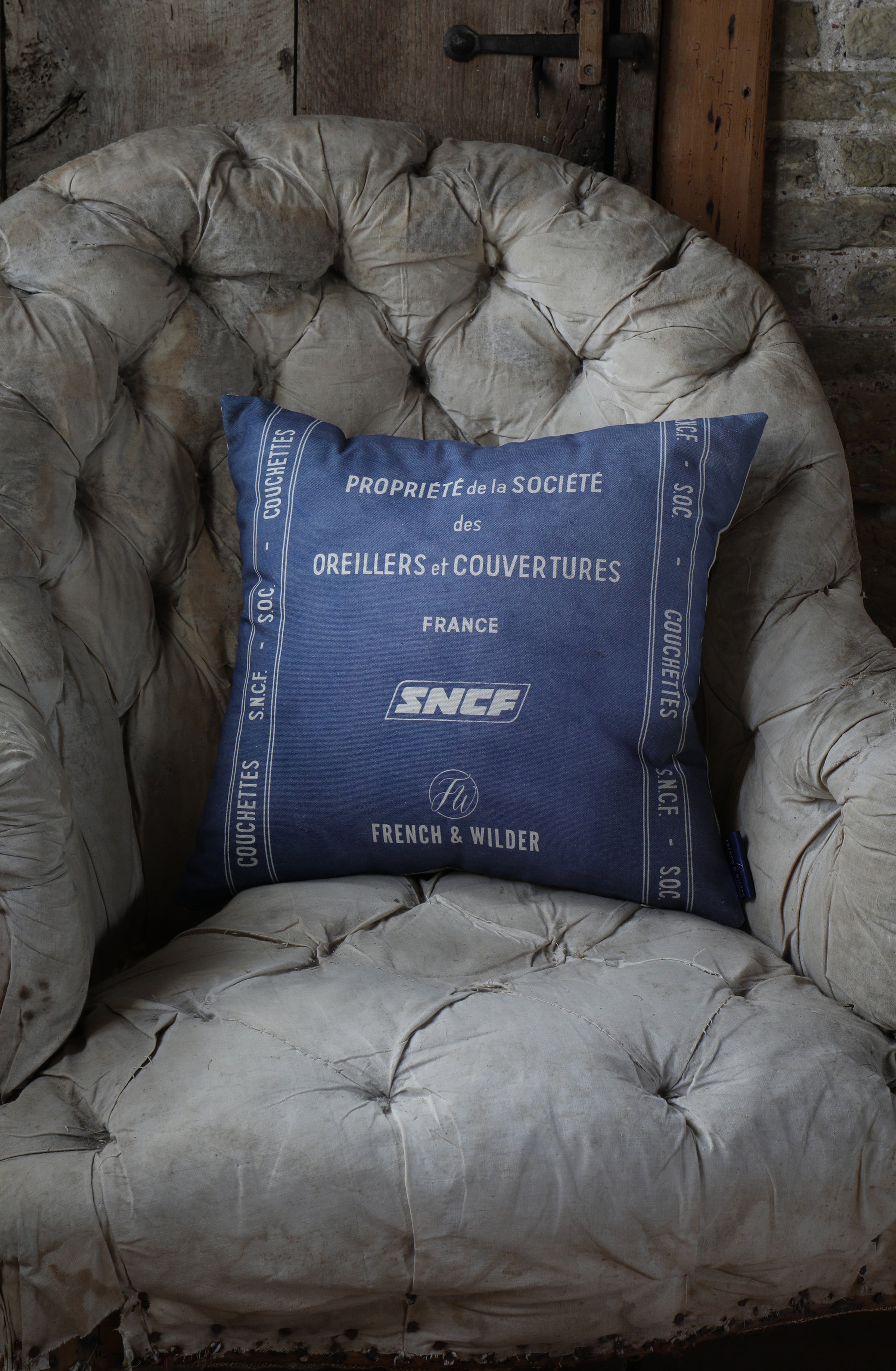 Cushion with blue and white SNCF design placed on a shabby vintage chair against a brick wall.