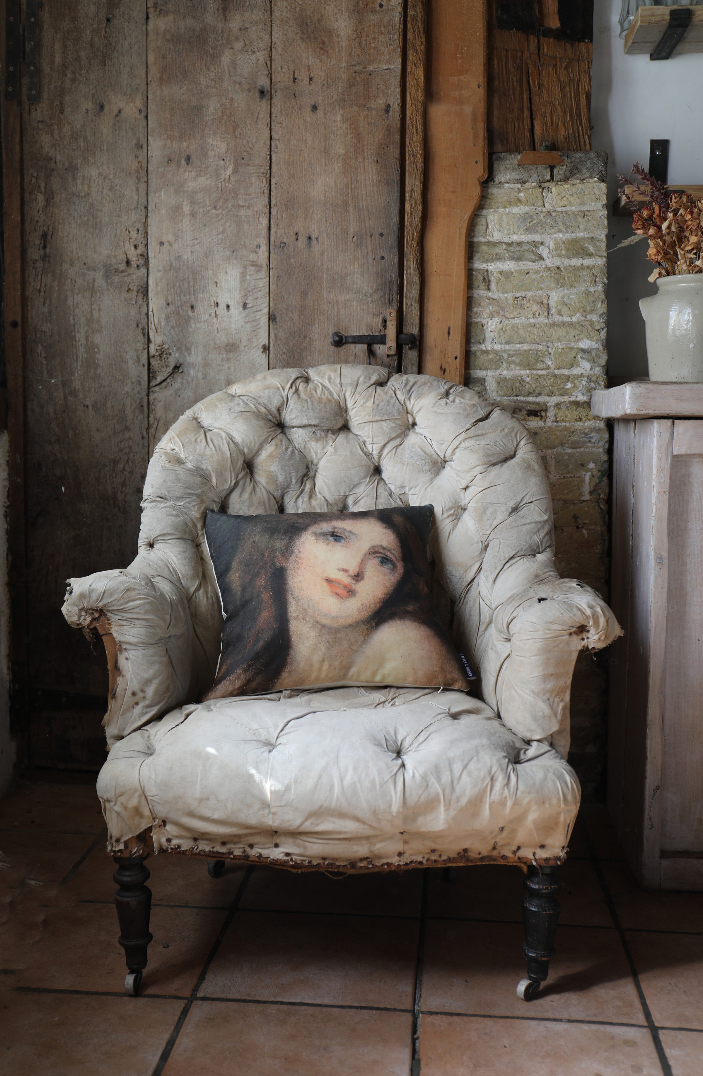 A cushion featuring a portrait of a woman in wistful pose placed on a vintage armchair in a rustic room setting