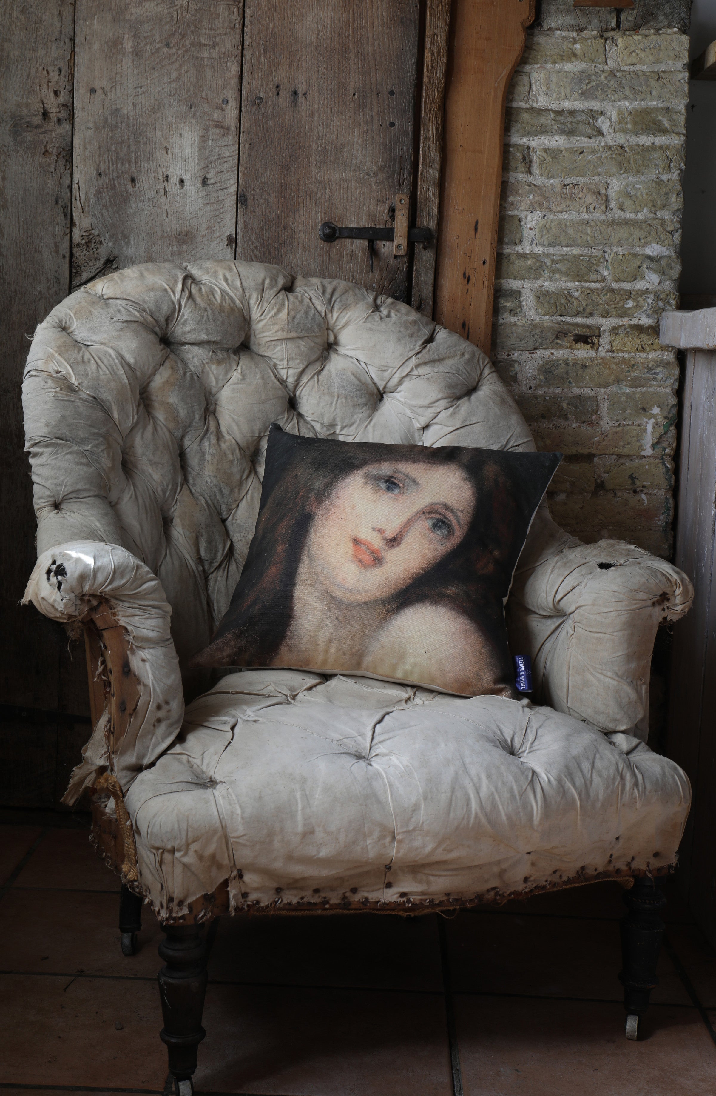 Decorative cushion with a woman's portrait design placed on a vintage armchair against a rustic brick wall.