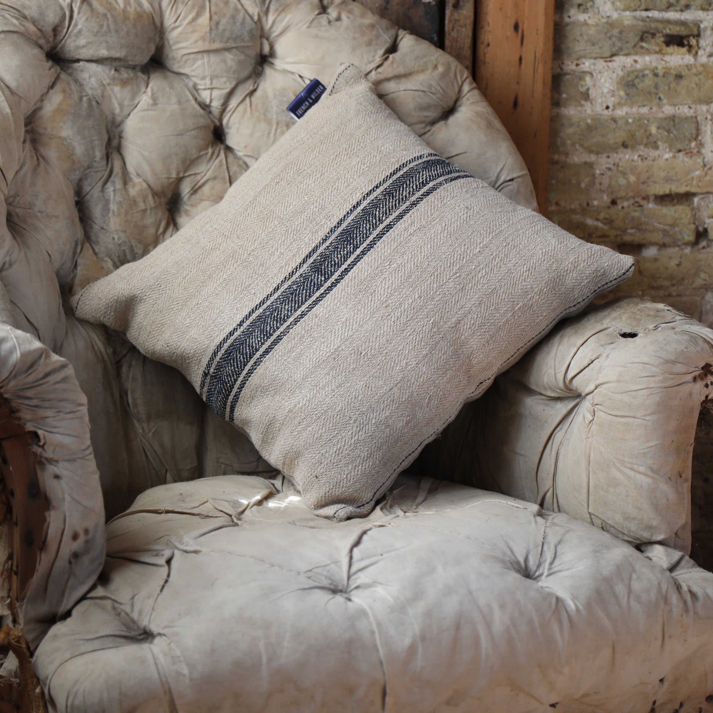 Cushion made from a vintage French sack hemp linen fabric placed on a worn armchair against a rustic brick wall.