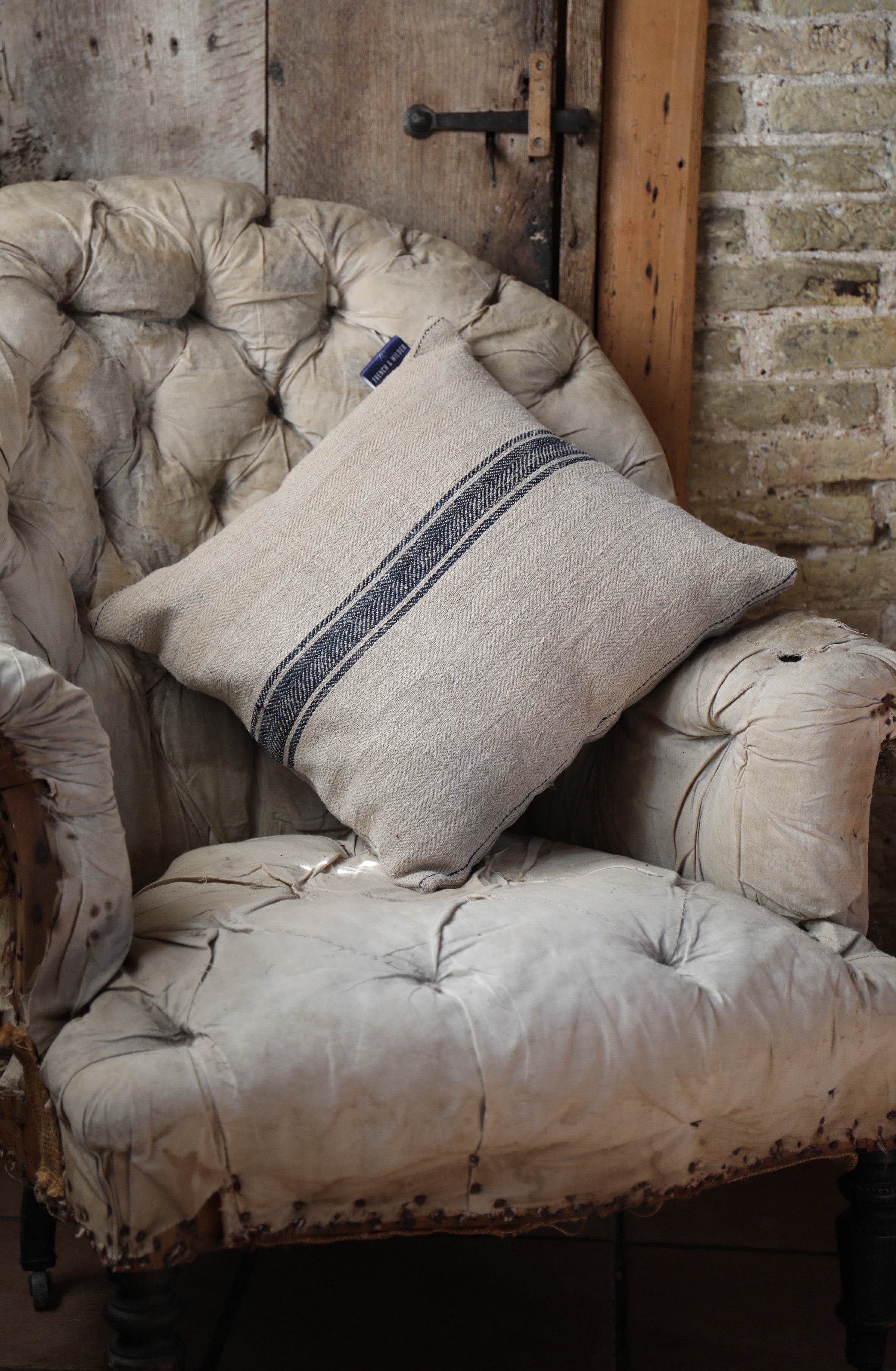 Cushion made from a vintage French sack hemp linen fabric placed on a worn armchair against a rustic brick wall.