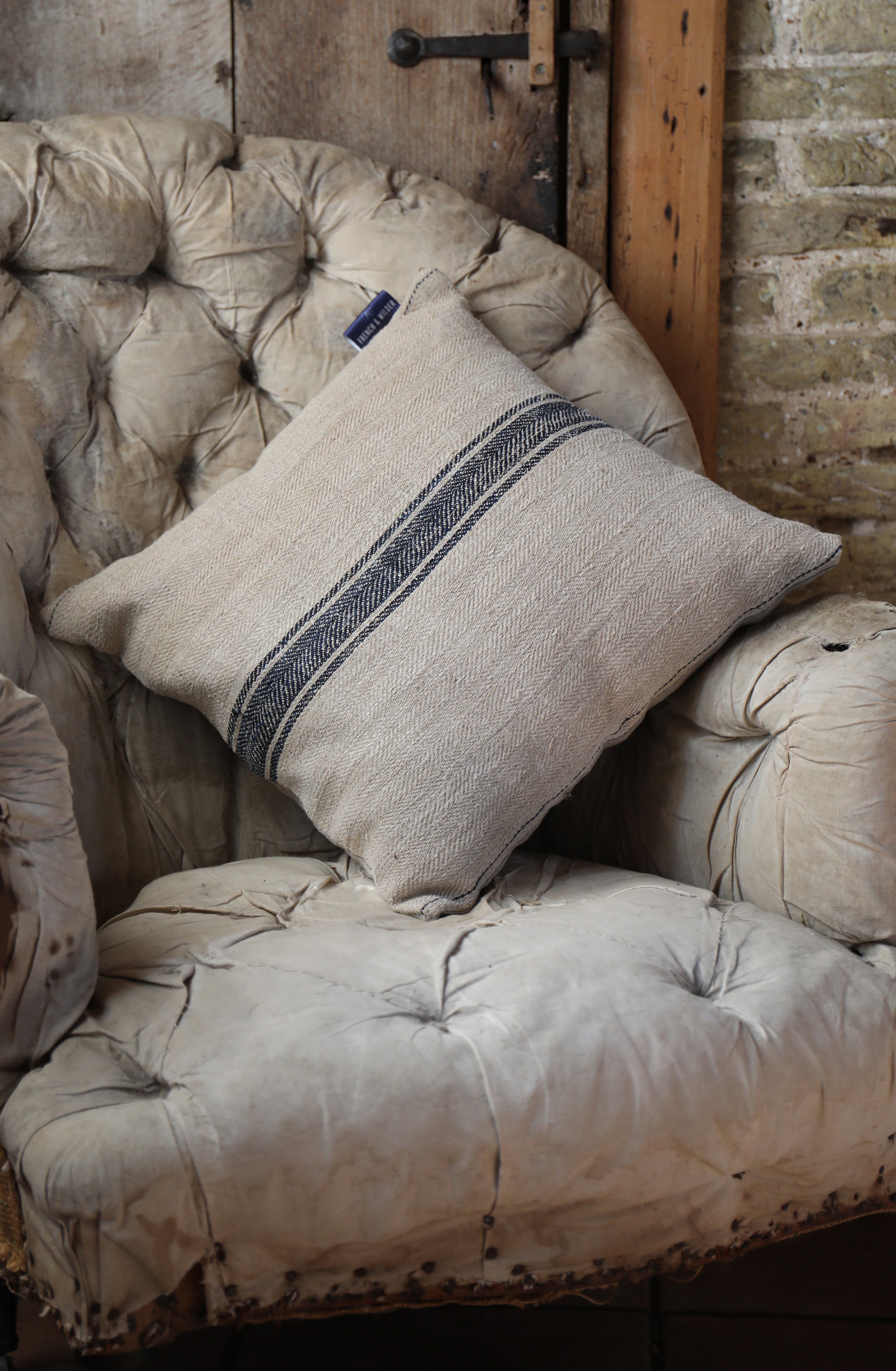 Cushion made from traditional sacking fabric, beige with dark blue stripe, sitting on an antique chair against a rustic brick wall.