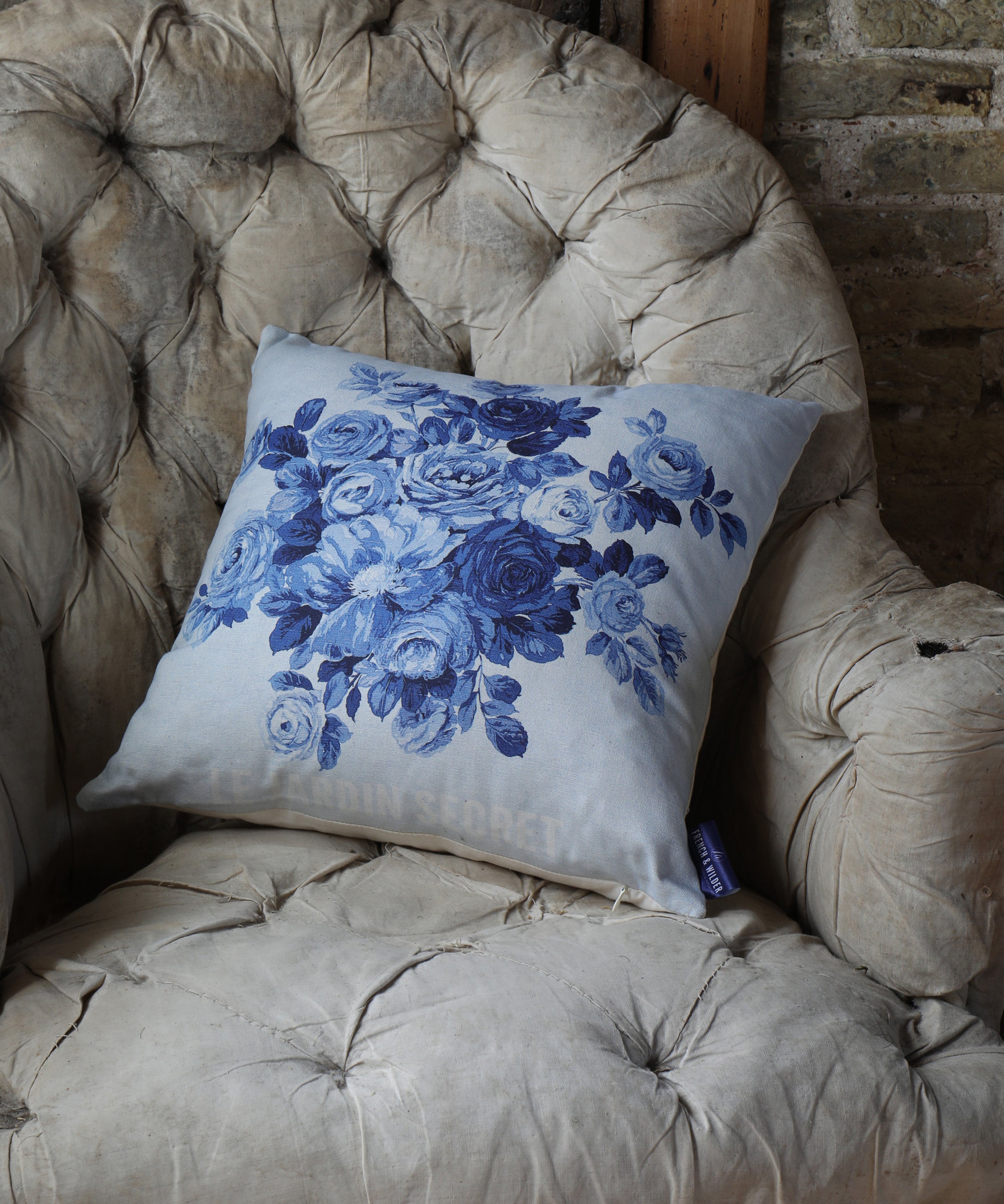 Cushion with blue floral pattern and the words 'Le Jardin Secret' on a beige vintage armchair against a brick wall.