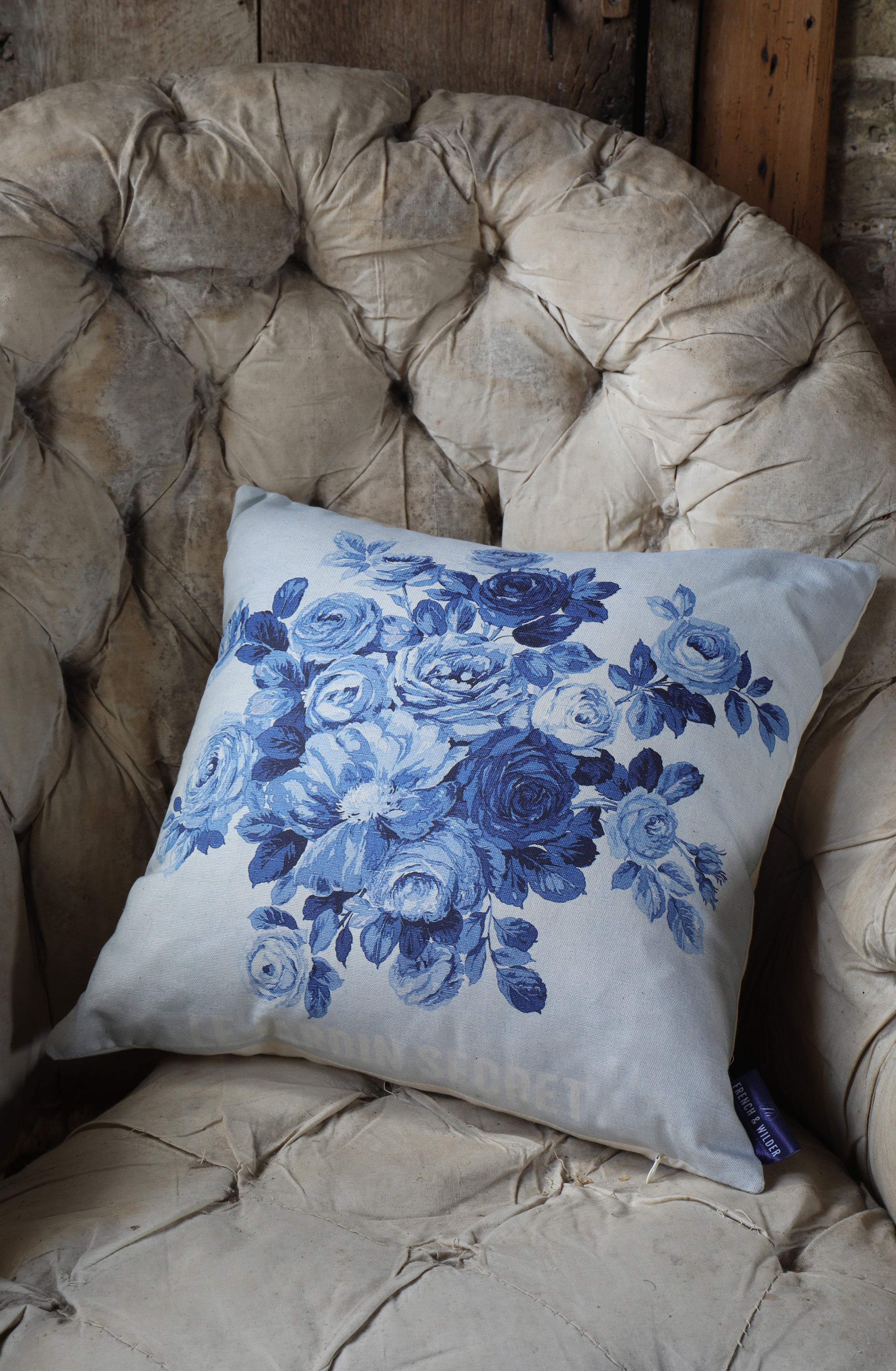 Blue floral pillow with roses and the text 'Le Jardin Secret' placed on a beige armchair.