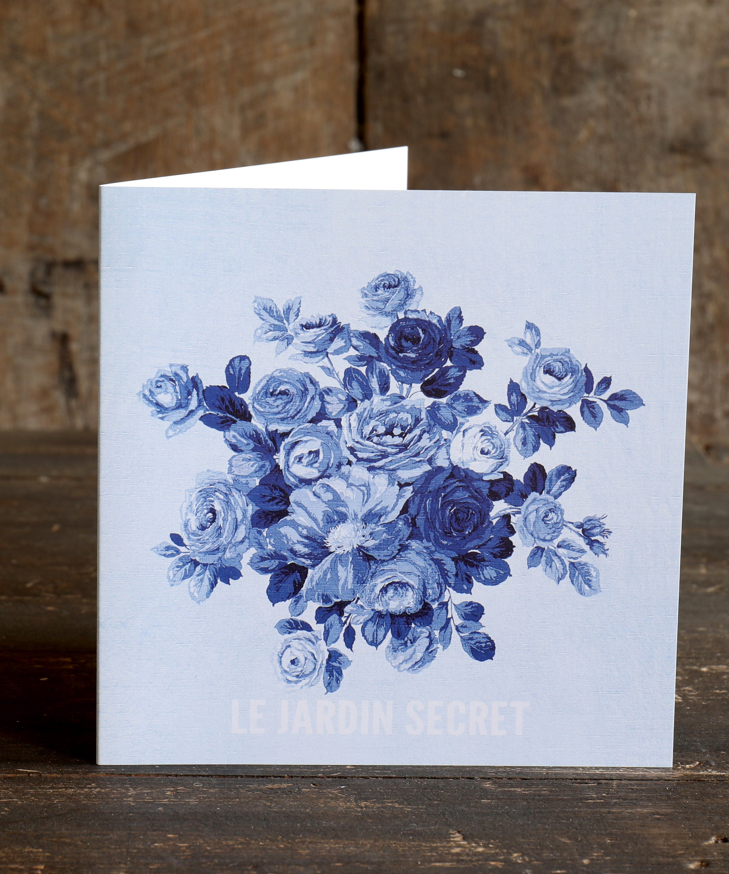 Greeting card with blue floral design of roses with the words 'Le Jardin Secret' below, on a wooden surface