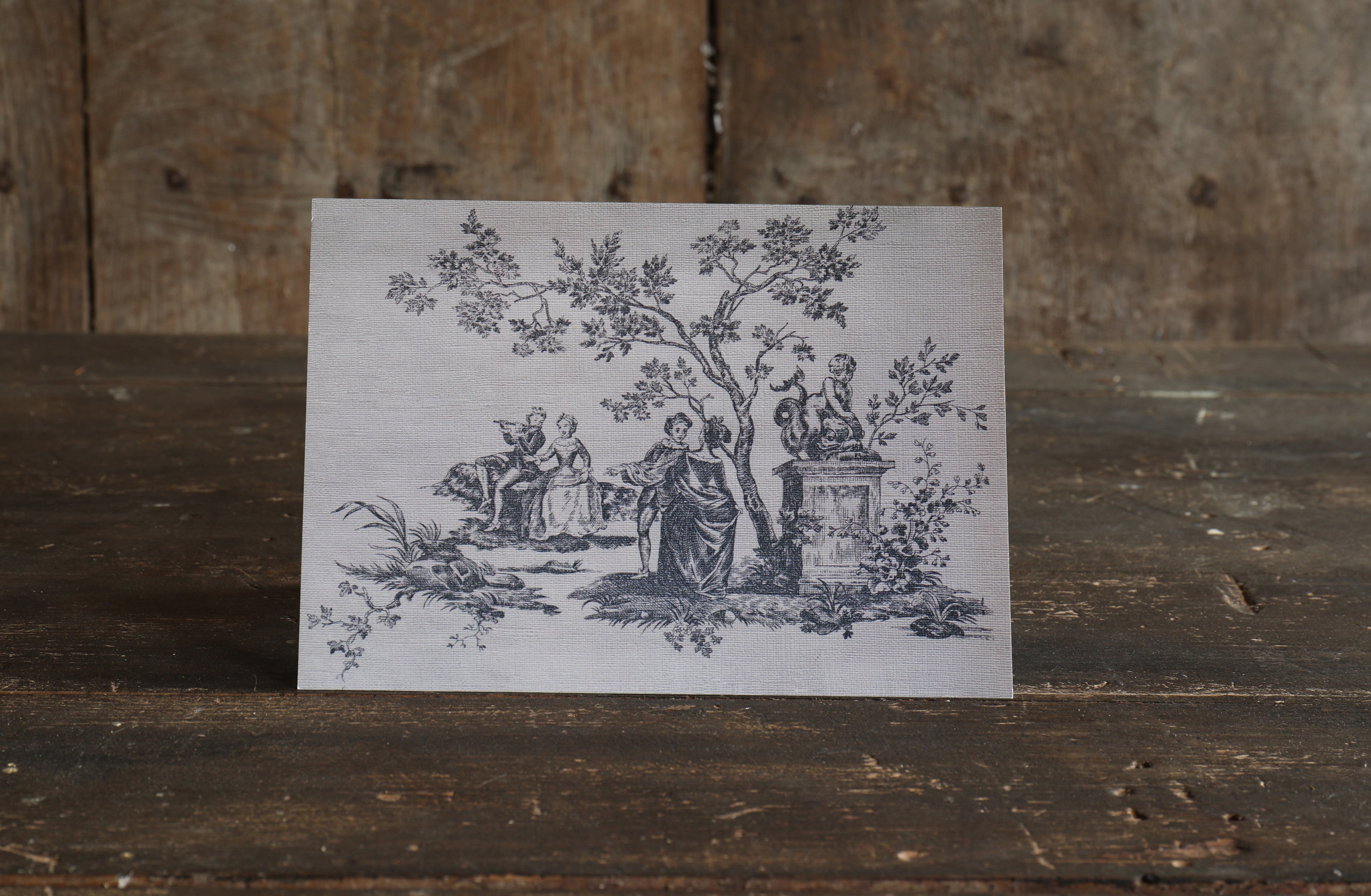 Greetings card with a textured vintage toile de jouy design of an illustration of people around a tree on a wooden surface.