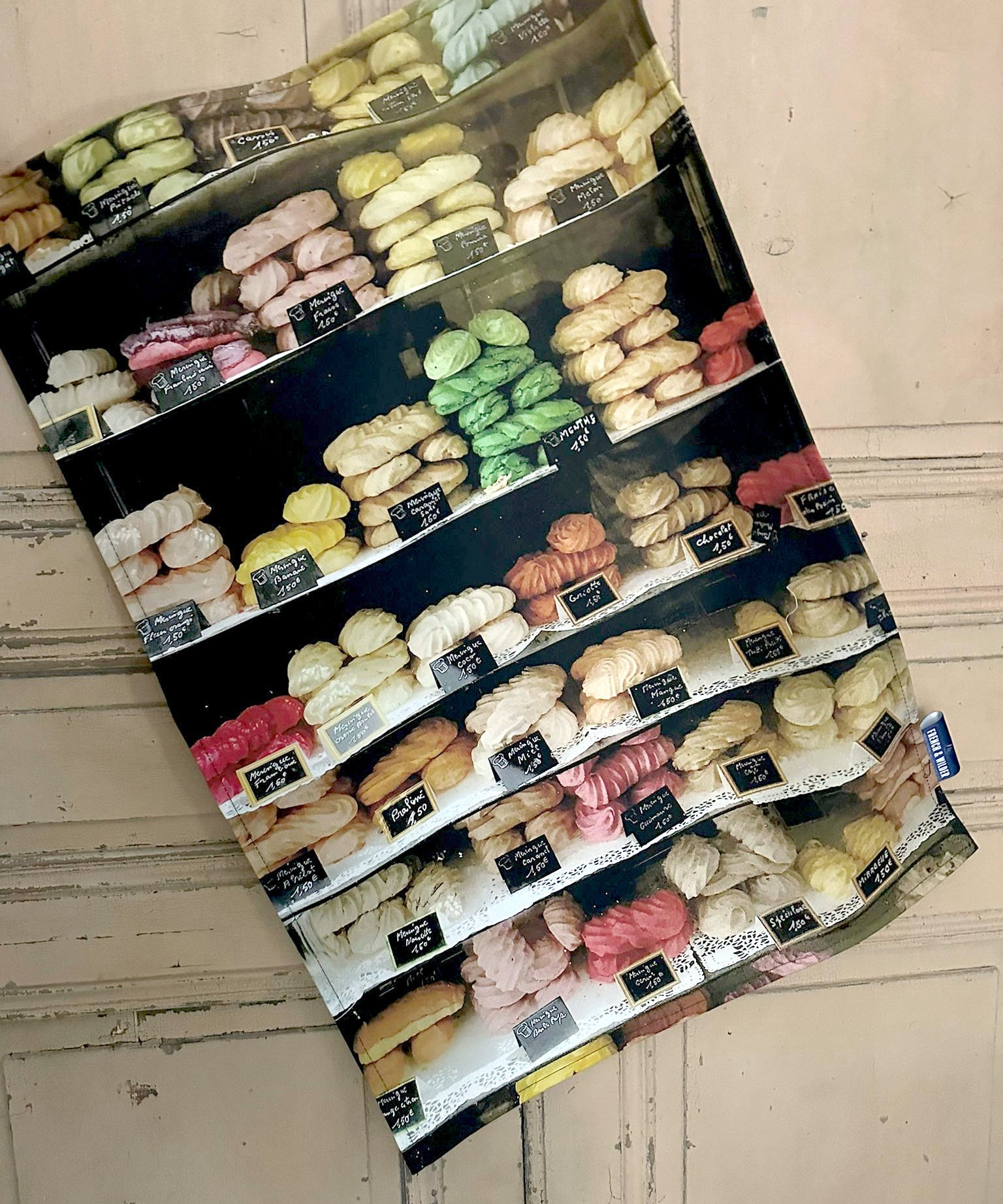 Tea towel featuring brightly colour selection of meringues in a French shop window hanging next to a light colour painted wooden door
