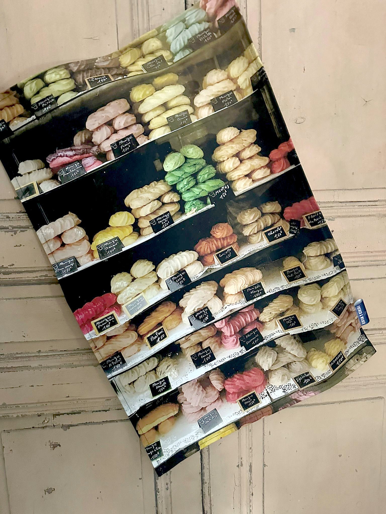 Tea towel featuring brightly colour selection of meringues in a French shop window hanging next to a light colour painted wooden door