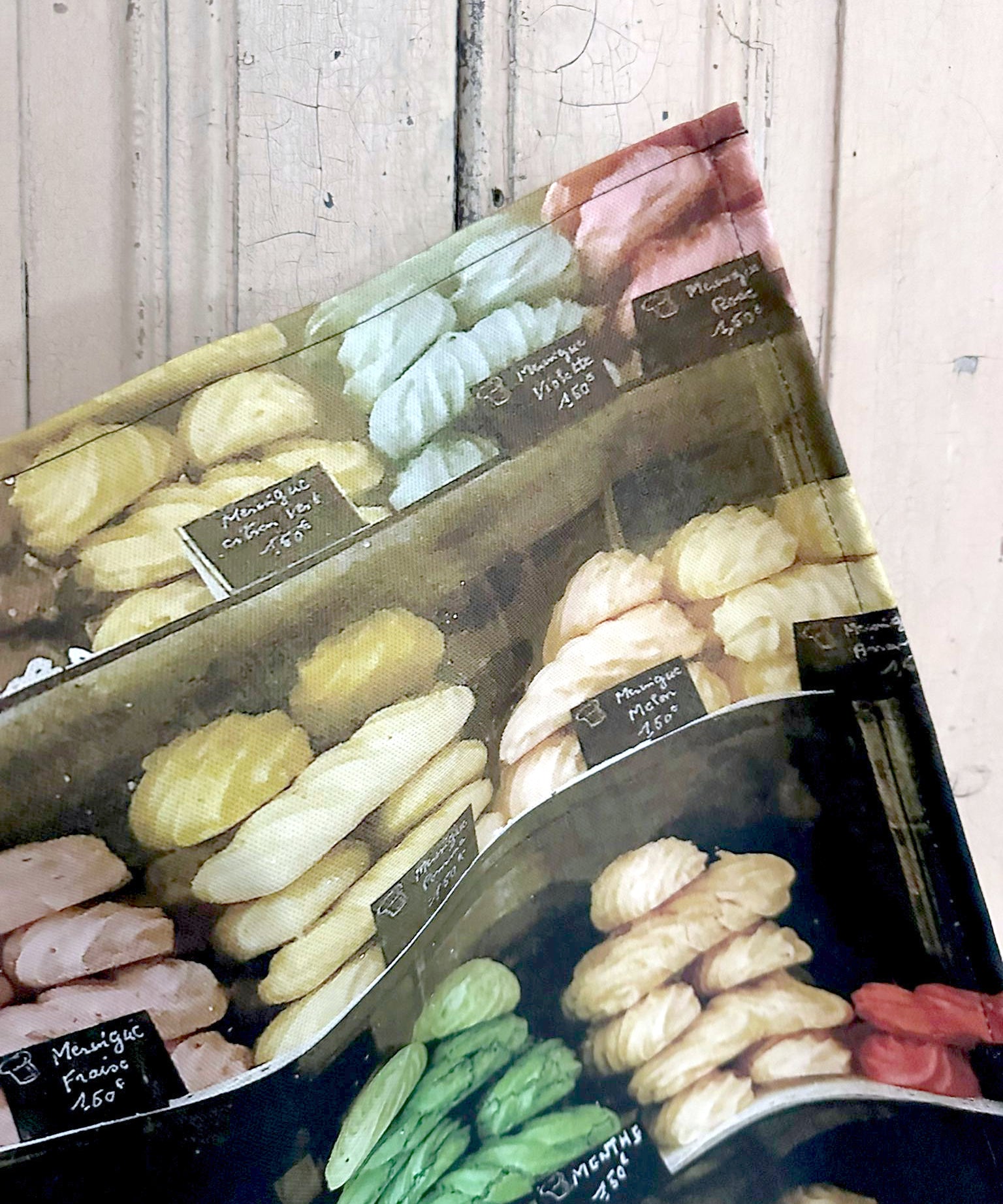 Detail of a tea towel featuring an image of meringues in a French shop window against a painted wooden surface.