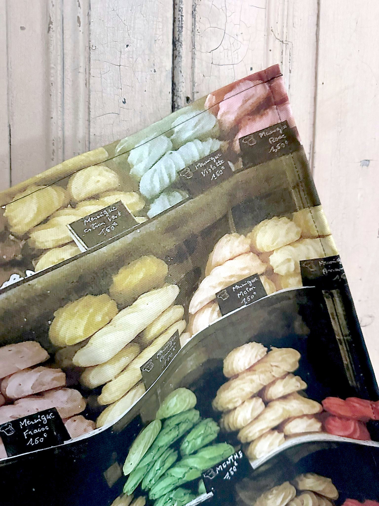 Detail of a tea towel featuring an image of meringues in a French shop window against a painted wooden surface.