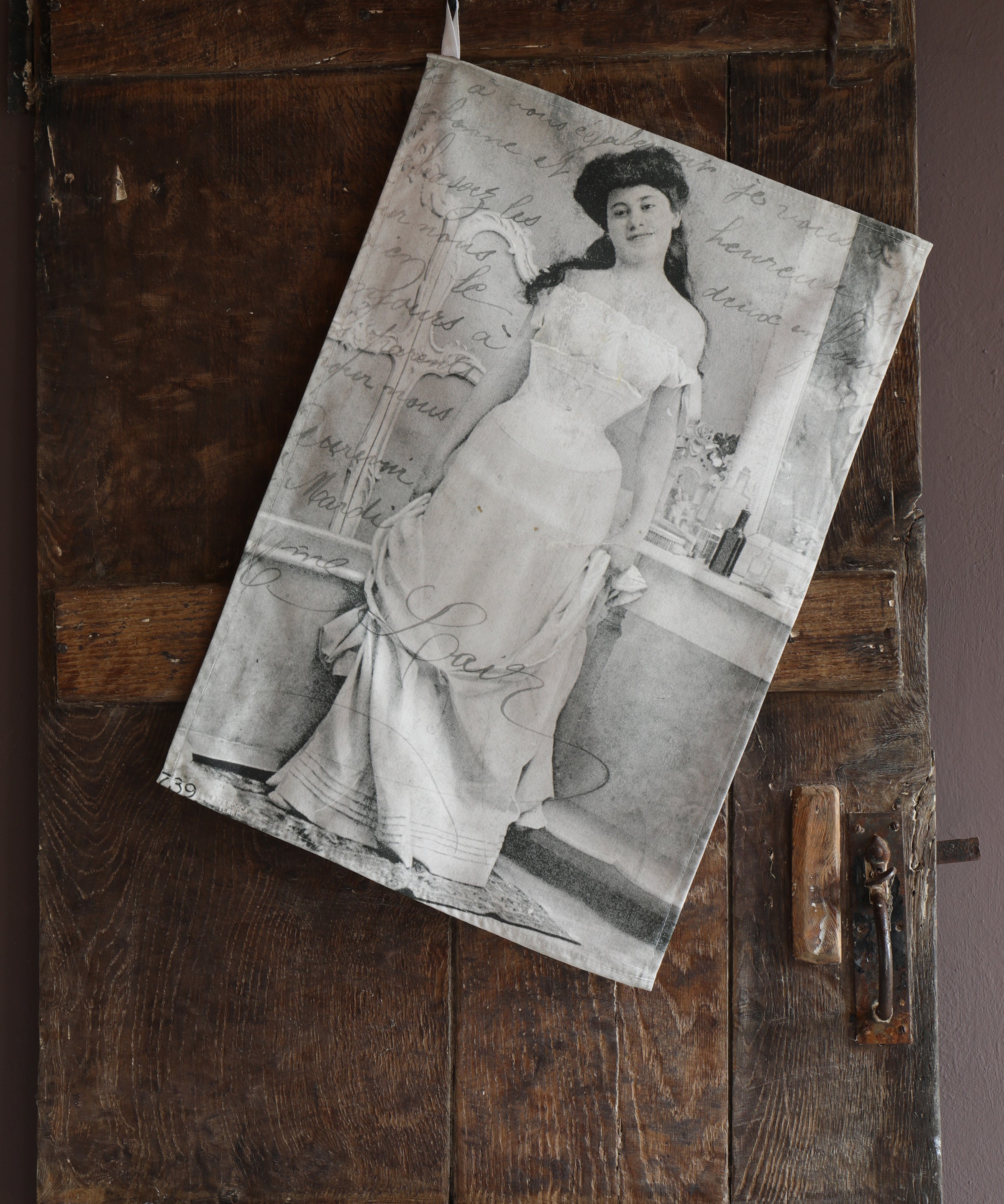 Tea towel hanging on an old wooden door with the image of a vintage postcard containing a black and white photograph of a smartly dressed woman