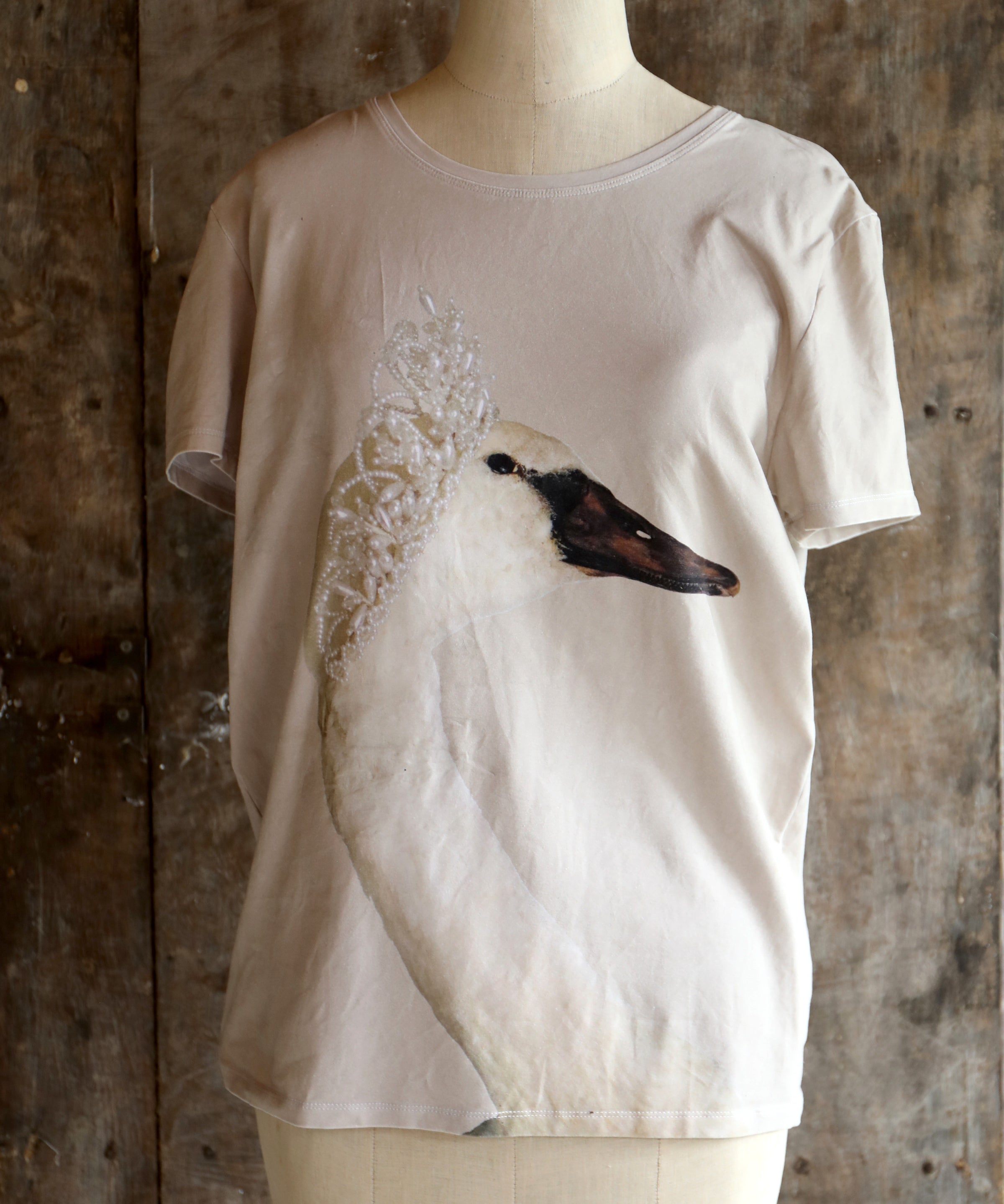 Beige t-shirt with a swan design on a mannequin against a textured wall.