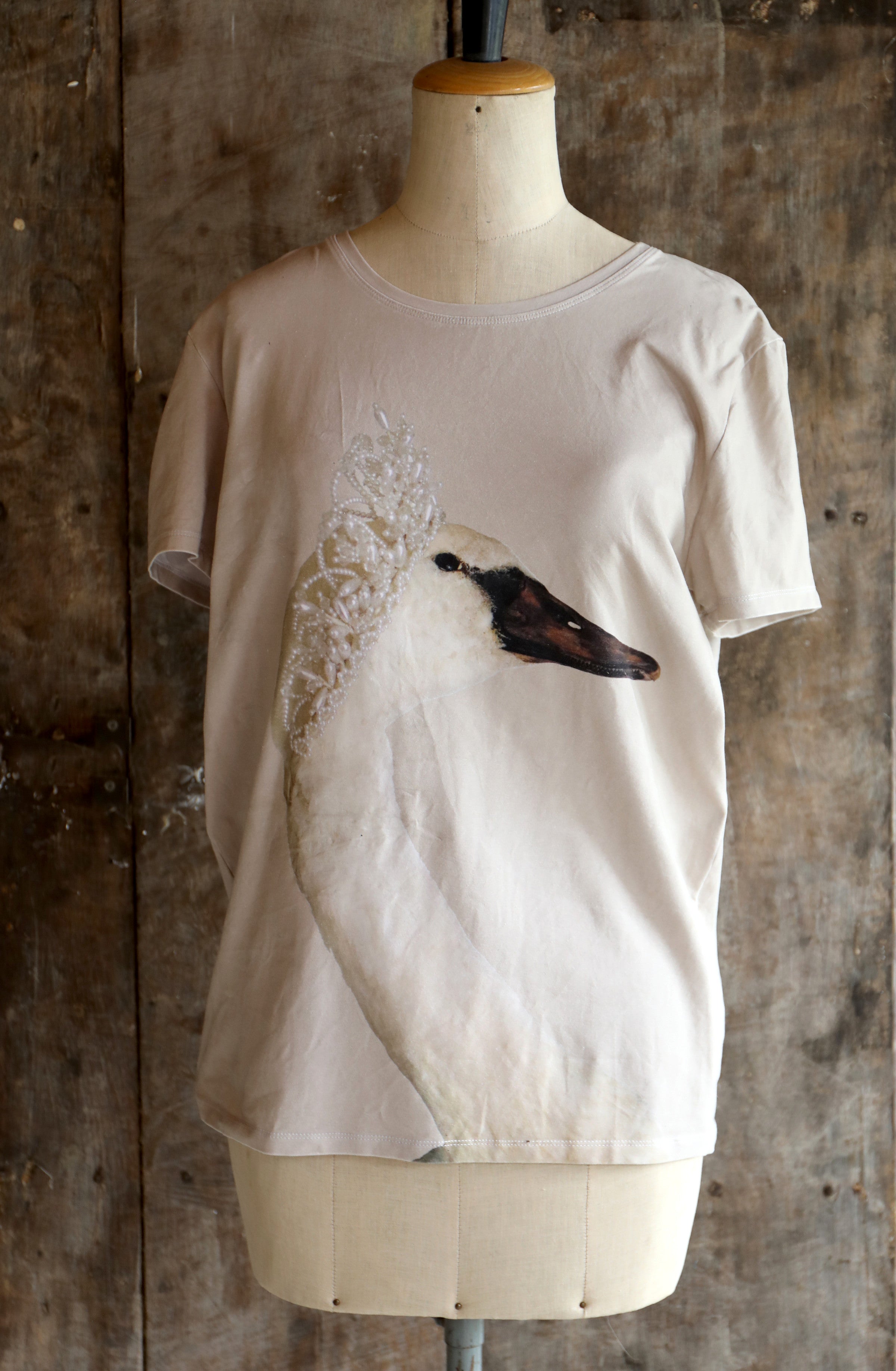 Beige t-shirt with a swan design on a mannequin against a textured wall.