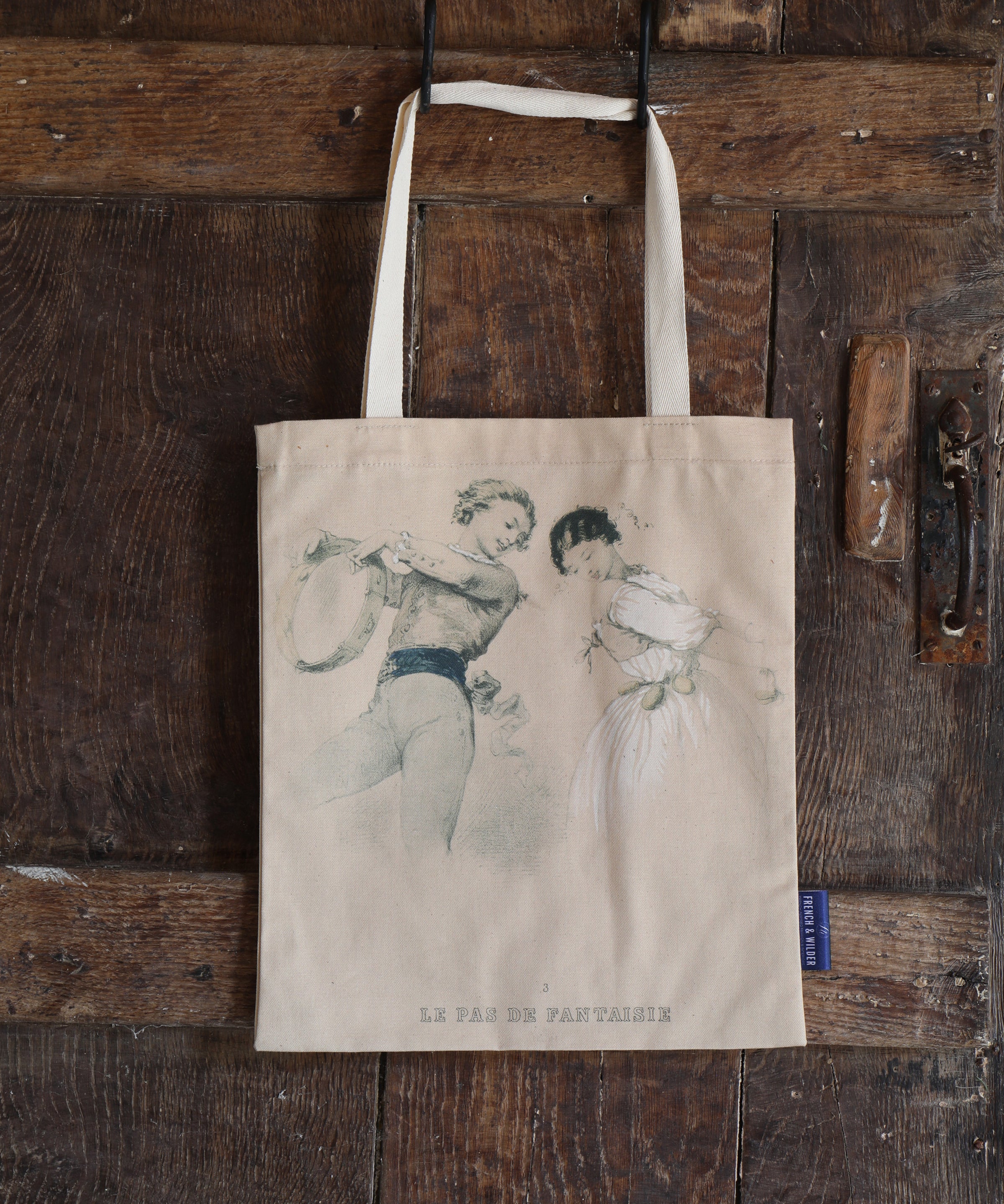 Tote bag hanging on an old wooden door with a line drawn vintage image of two young lovers with the subtitle 'Le Pas de Fantaisie'.