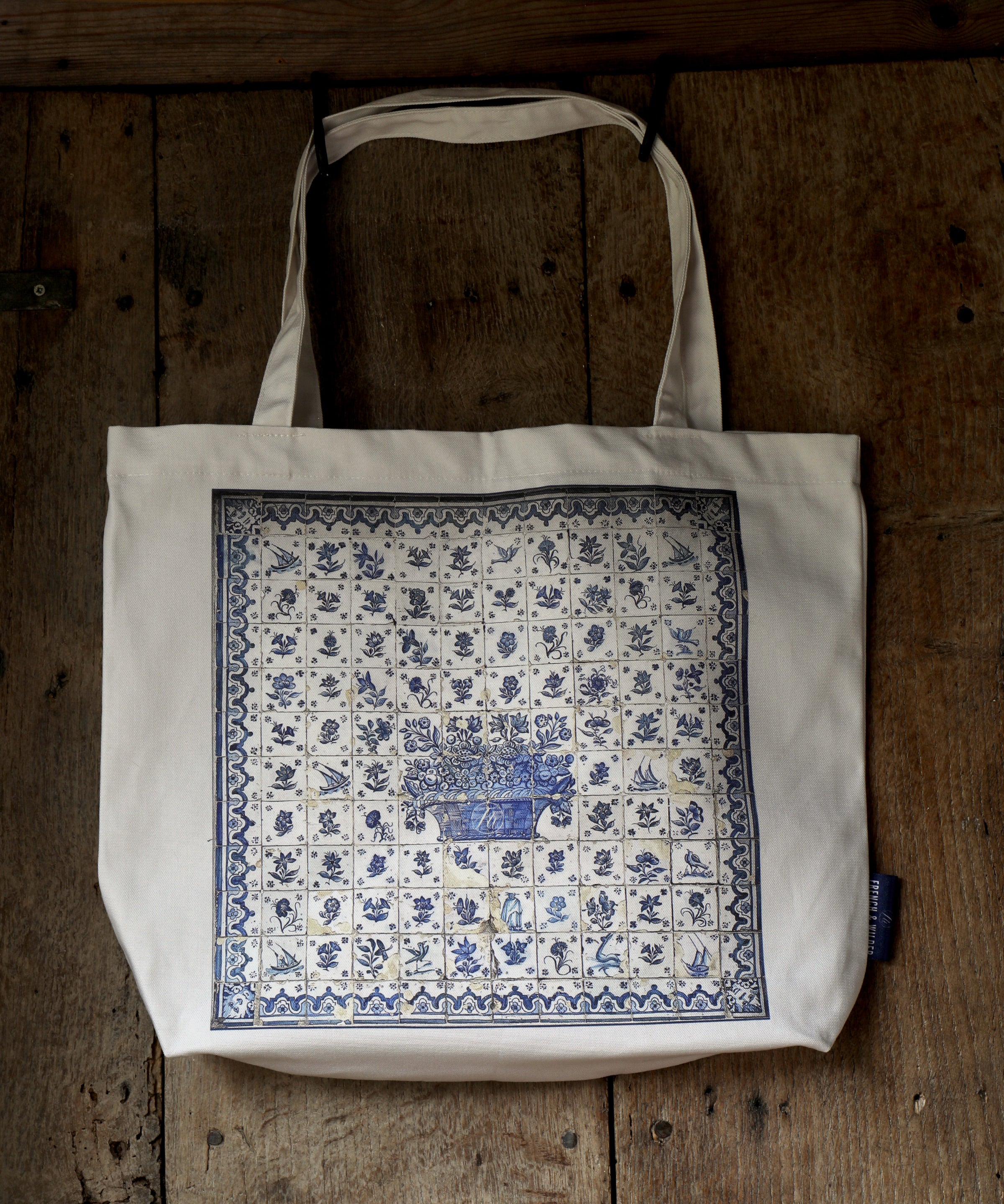 Pale coloured tote bag with blue tile pattern design hanging from an old rustic wooden door