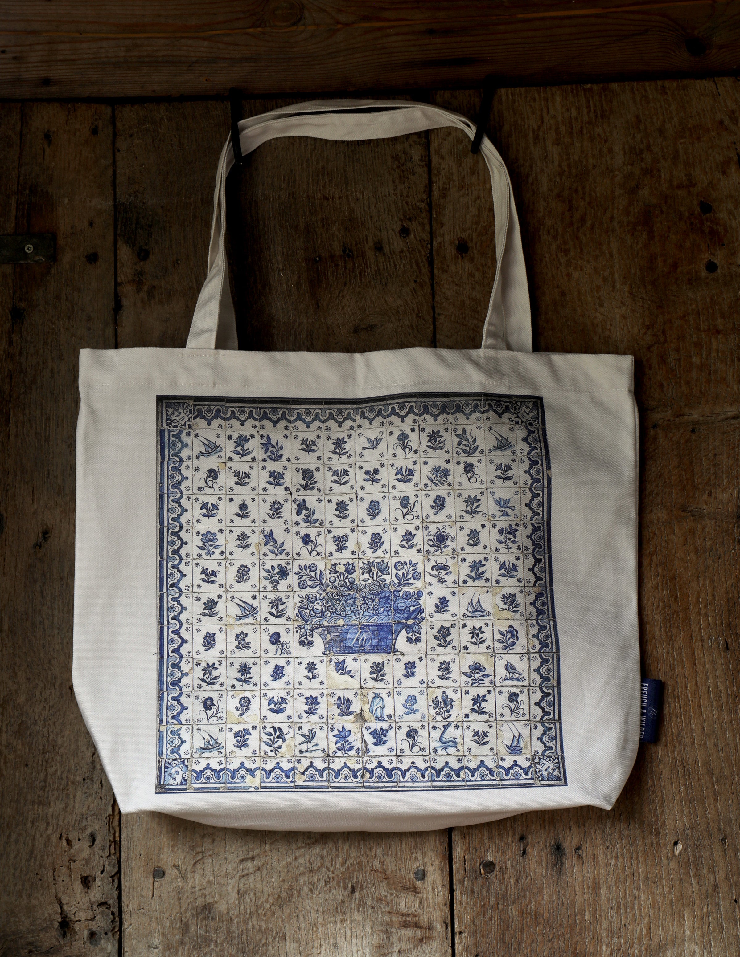 Pale coloured tote bag with blue tile pattern design hanging from an old rustic wooden door