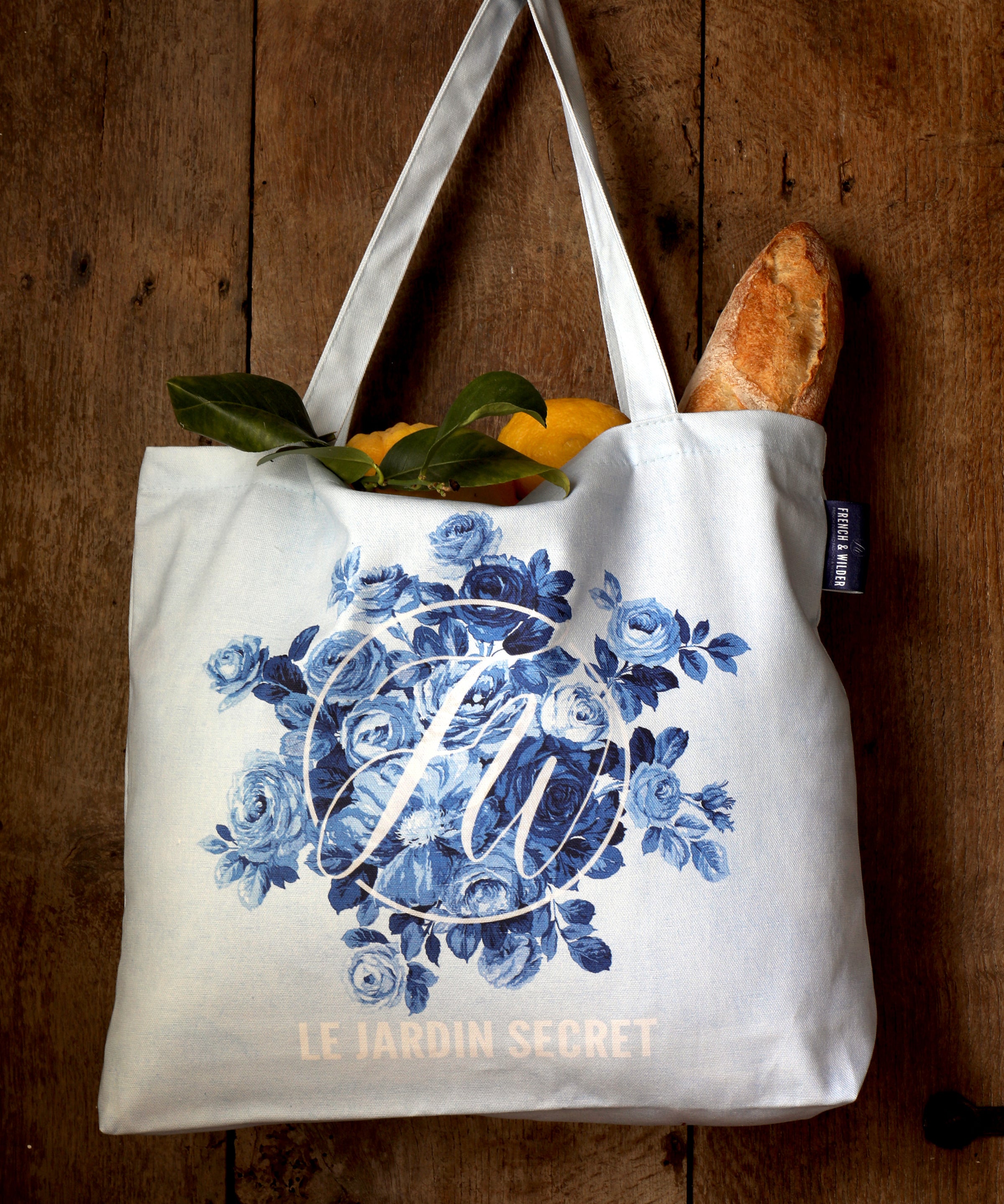 Tote bag with blue floral design, 'Le Jardin Secret' text and FW monogram hanging from a hook on an antique wooden door