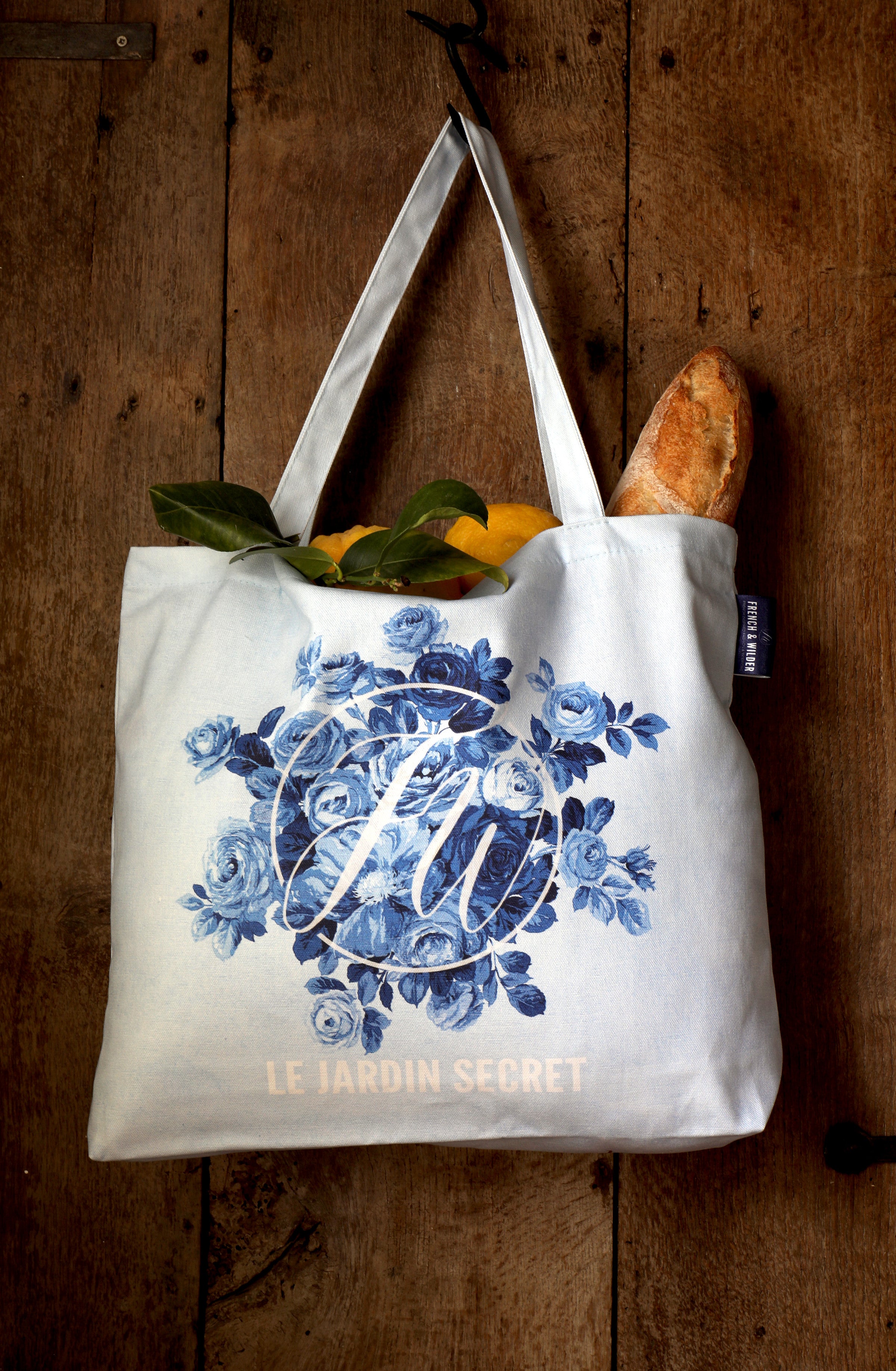 Tote bag with blue floral design, 'Le Jardin Secret' text and FW monogram hanging from a hook on an antique wooden door