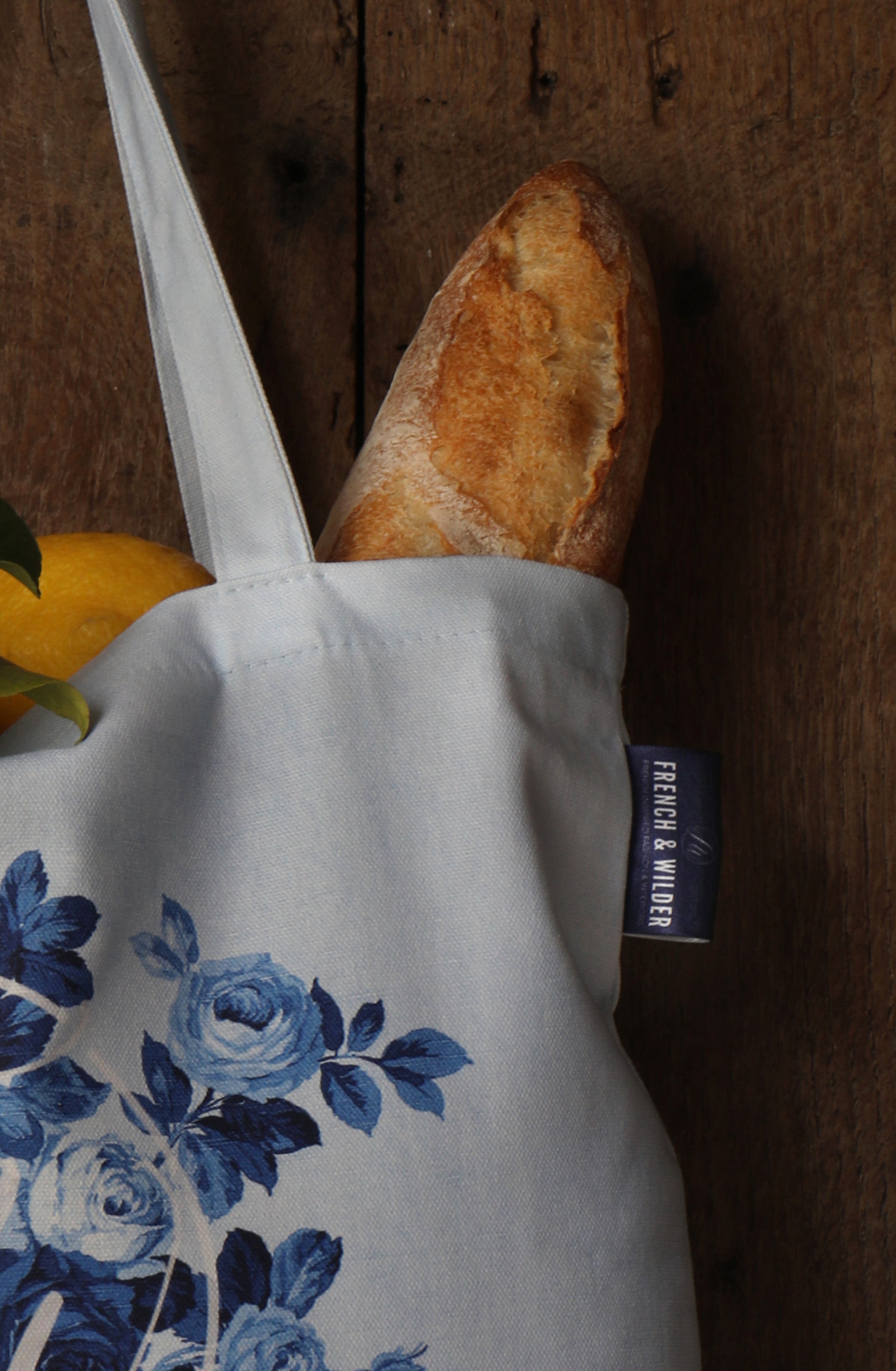 A close-up view of a blue floral pattern tote bag containing a baguette and lemons next to a wooden surface