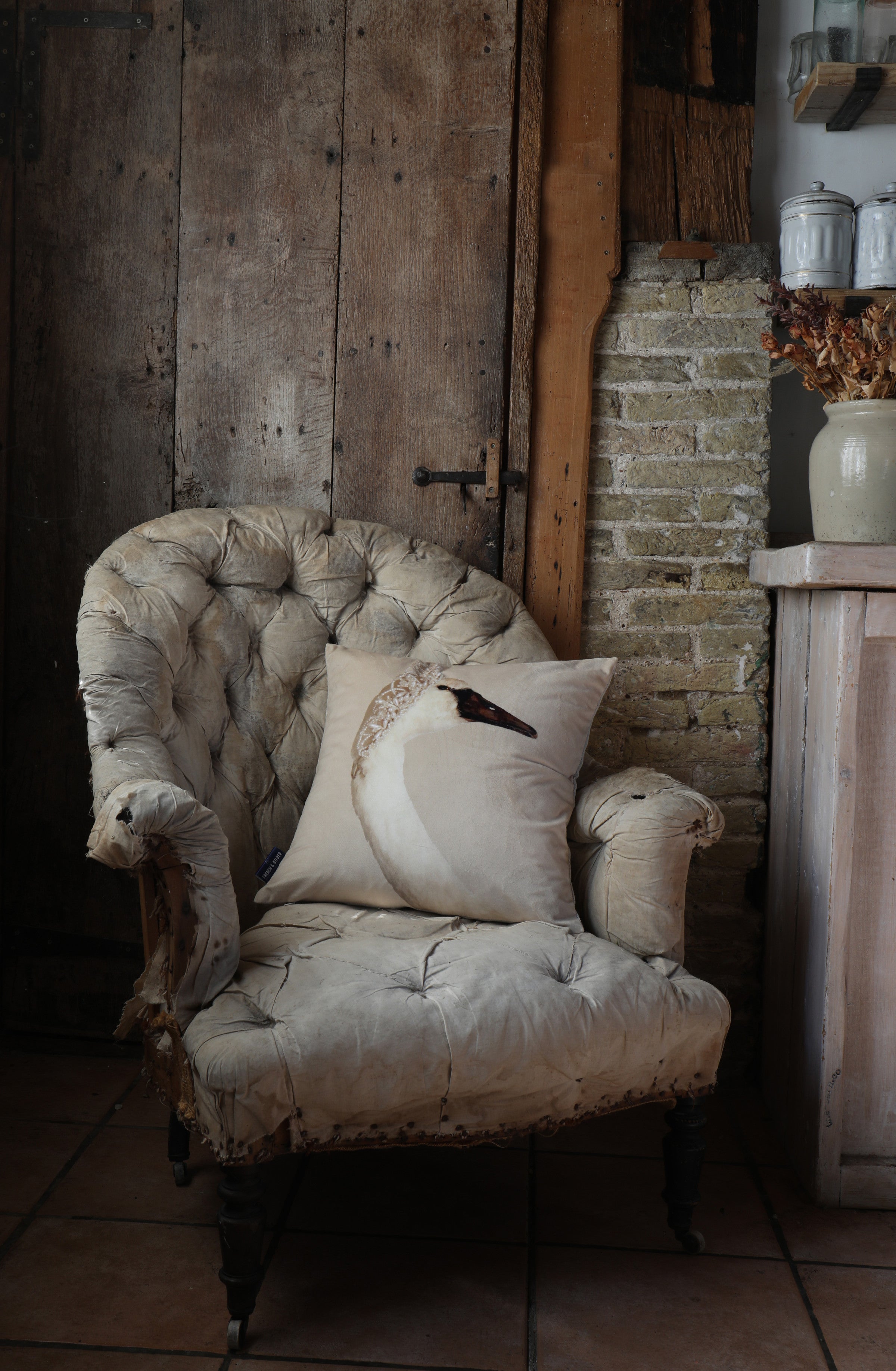 Descorative cushion featuring a swan wearing a tiara, placed on a vintage armchair in a rustic room with a brick wall and old wooden door
