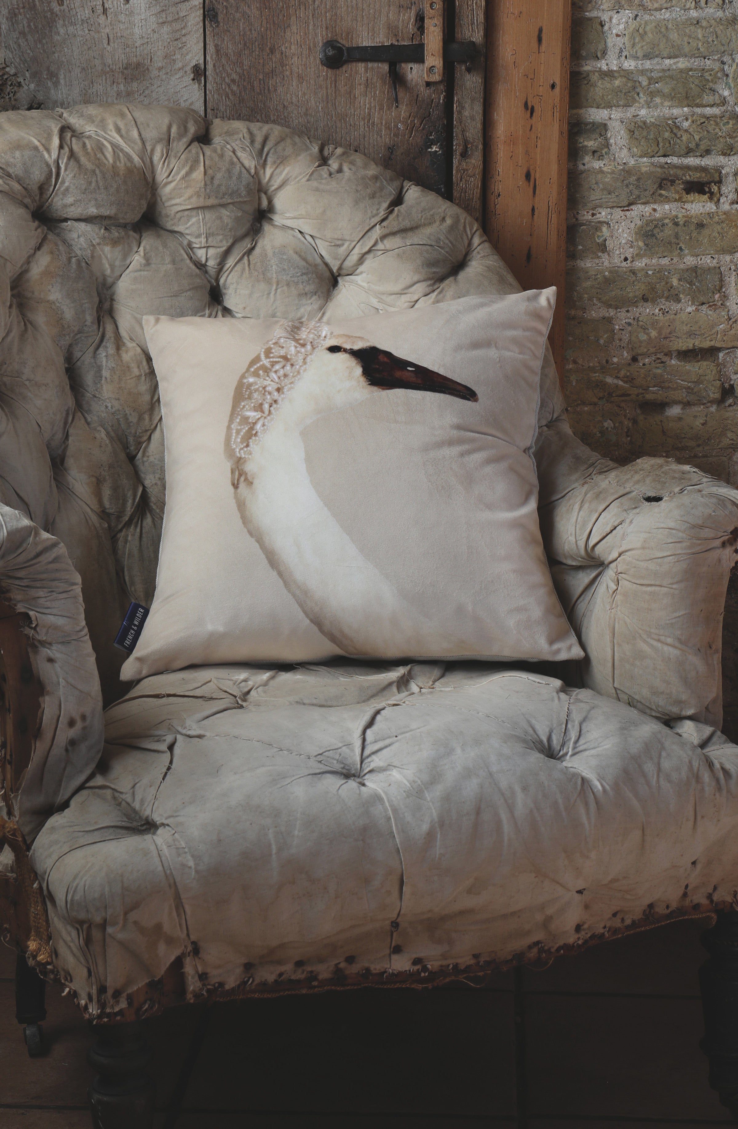 Decorative pillow with a swan design on a vintage chair against an old wooden door and a rustic brick wall.